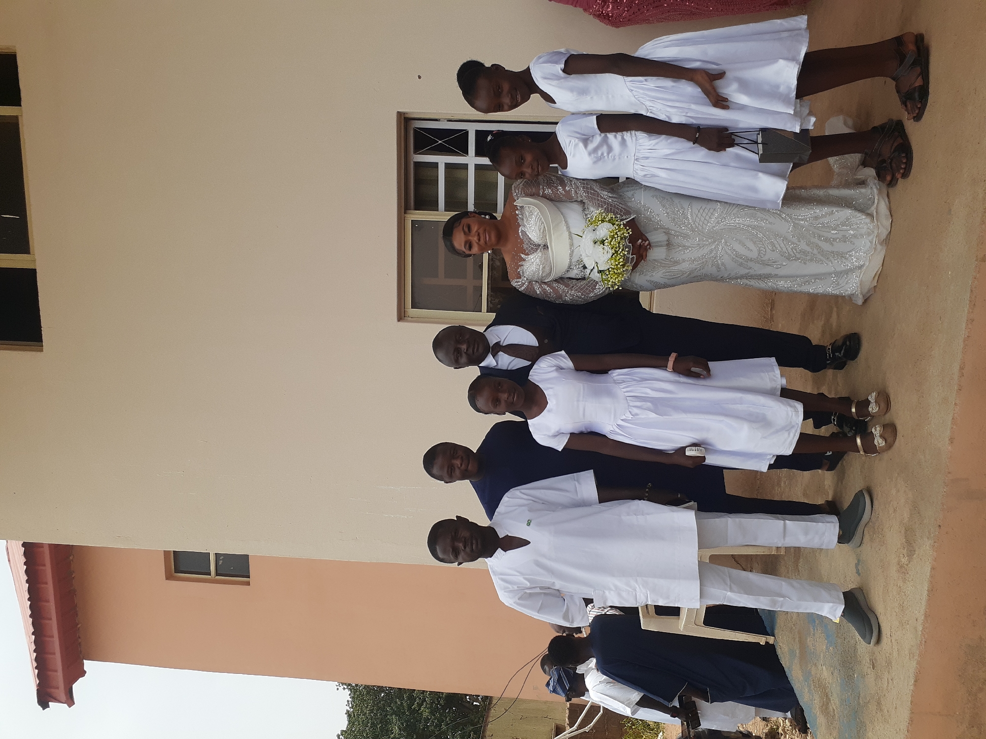 A wedding group photo taken outdoors in Barakin Lerawi, Nigeria. A bride in a silver, long-sleeved gown stands with a bouquet in front of a window. Two flower girls in white dresses stand to her right. A groom in a black suit and tie stands next to the bride on her left. Two groomsmen in dark blue suits stand next to the groom. A groomsman wearing a white suit stands to the left of the others. Behind the group is a beige building with a red roof.