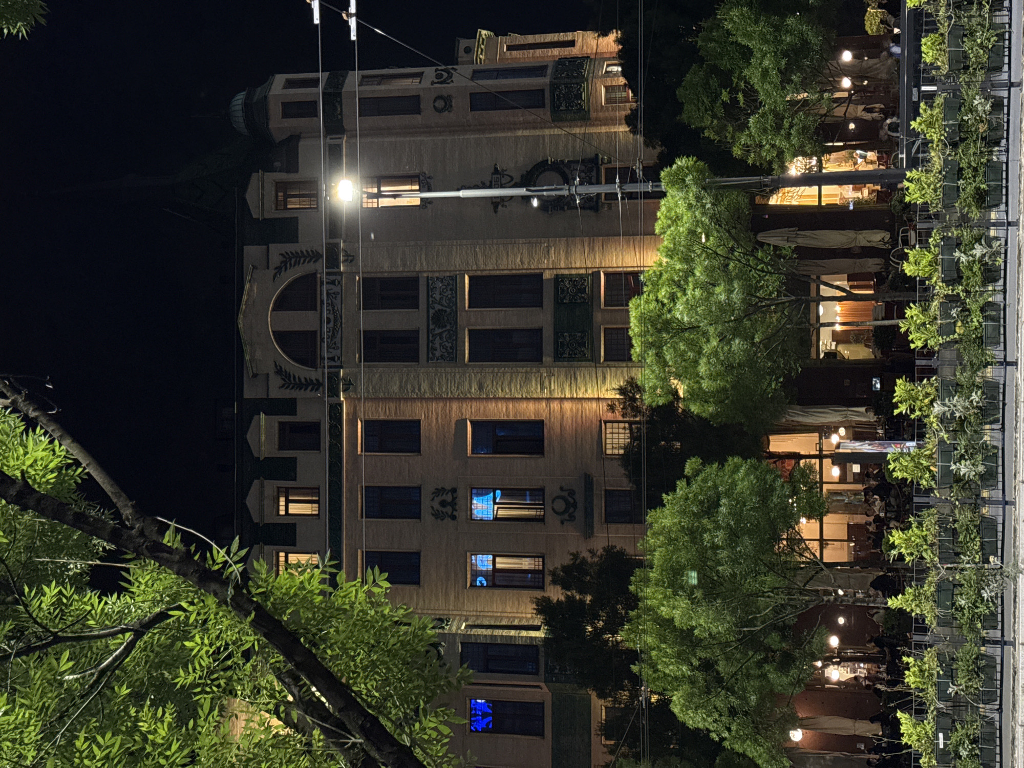 The image captures a nighttime scene in Belgrade, Serbia. The architectural focus is a tall, ornately decorated building with illuminated windows. A row of trees and outdoor dining umbrellas partially obscures a restaurant, where people are seated. Street lighting casts a glow on the scene.