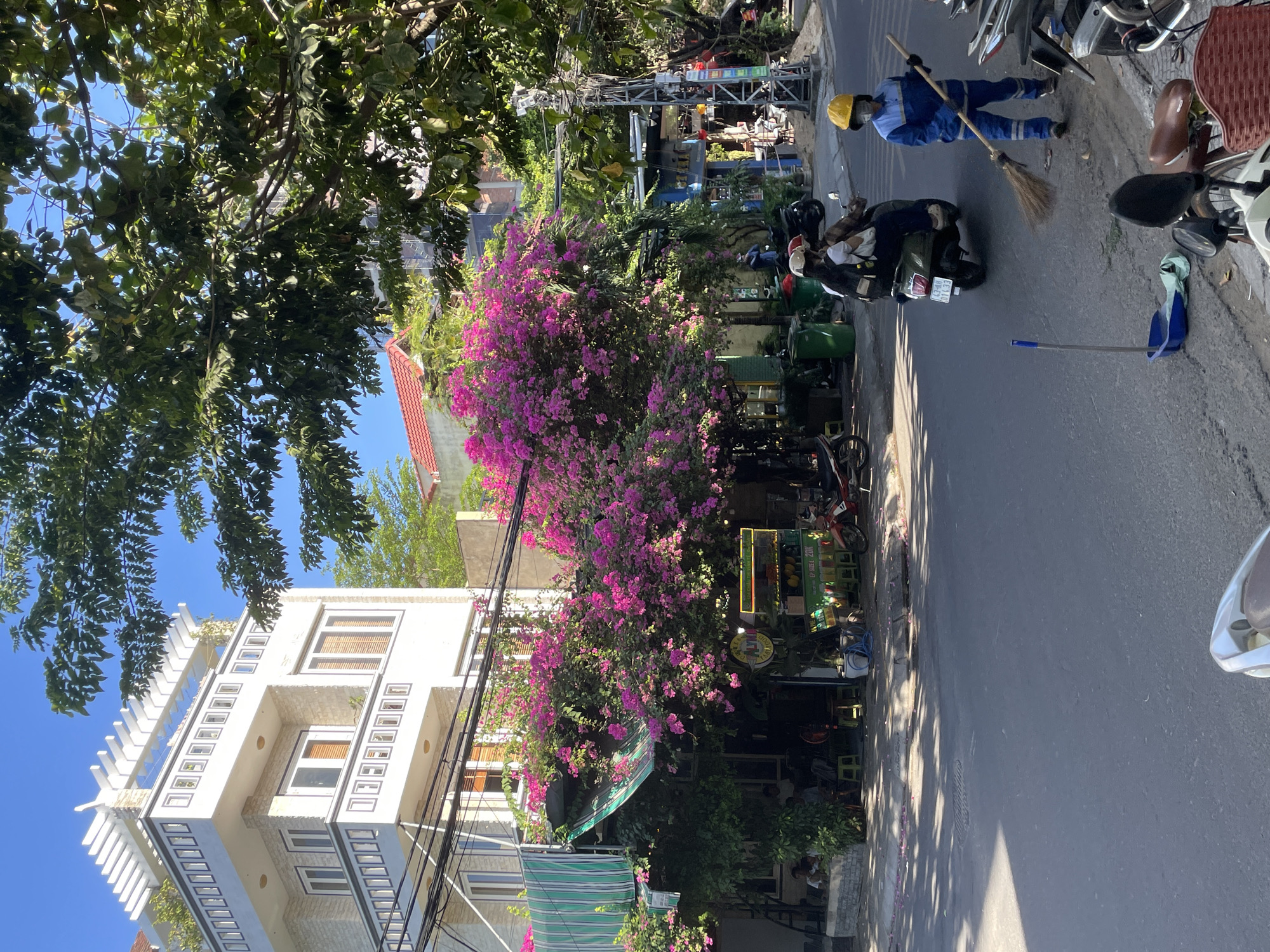 The image depicts a street scene in Đà Nẵng, Vietnam, on a sunny day. In the foreground, a sanitation worker wearing a blue uniform and a yellow hard hat is sweeping the sidewalk with a broom. Next to them is a blue dustpan containing some debris. Several motorcycles are parked on the side of the street. Further down the street, a green scooter with two passengers is moving away from the camera.
Behind the sidewalk, a building with a white facade and multiple balconies is visible. Lush pink bougainvillea flowers spill over a green awning, covering part of the building. Under the awning, there appears to be an outdoor seating area, with people visible sitting at tables. Next to this establishment, a green cart with colorful displays, possibly selling fruit or drinks, is set up. Power lines crisscross the scene, and a tall metal utility pole stands on the right. The sky is clear and blue.