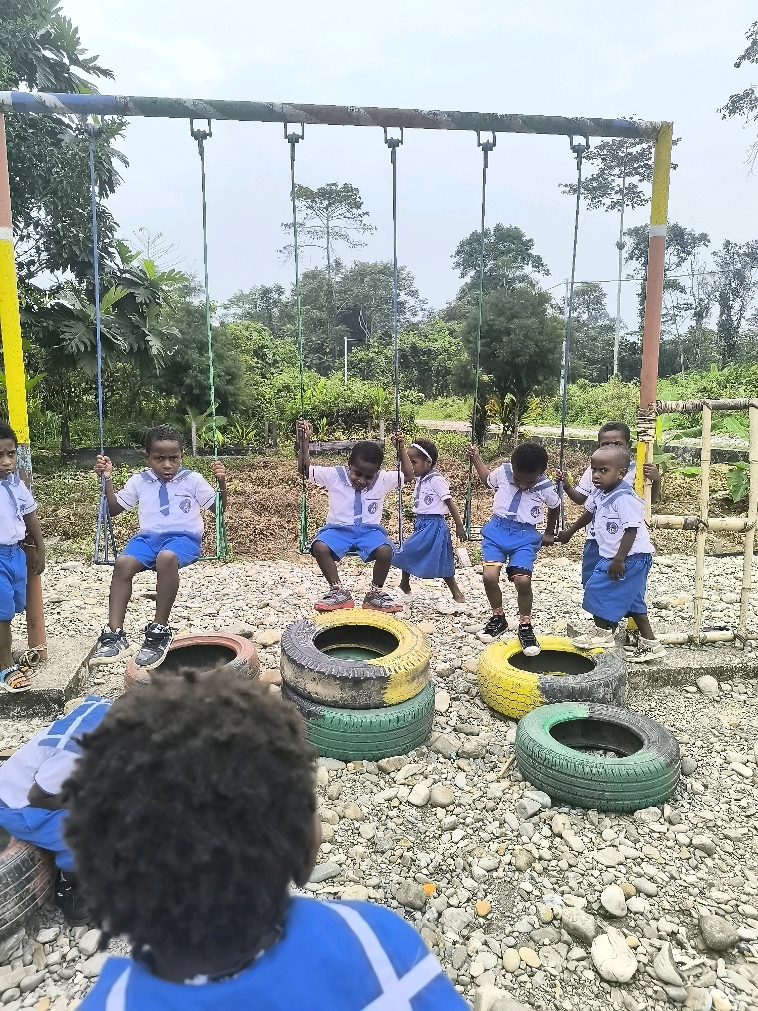 The image depicts a group of children playing on a swing set constructed from repurposed tires in Timika, Indonesia. The swing set is situated in an outdoor environment with a backdrop of lush green vegetation and trees, suggesting a rural or semi-rural setting on the outskirts of the city. The ground is covered with stones and several stacked tires serve as obstacles or seating areas around the swing set.
There are at least six children visible, all wearing school uniforms consisting of white shirts with blue ties and blue shorts or skirts. Some children are on the swings, actively playing, while others are standing or walking around. One child in the foreground is facing away from the camera, with their back to the viewer, their dark curly hair prominent. The children appear to be engaged in recreational activities, with expressions of enjoyment or concentration on their faces. The overall scene conveys a sense of childhood play and community engagement in an environment that utilizes readily available materials for recreation.