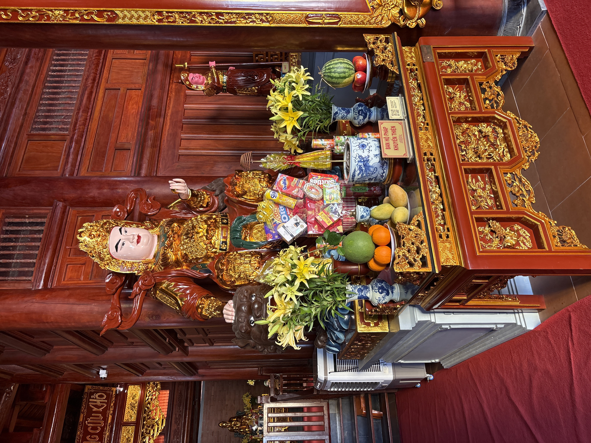 The image depicts a wooden altar in what appears to be a temple or religious space, potentially located in Nam Nha Trang Ward, Vietnam. A reclining Buddha statue, adorned in gold, lies on the altar. The altar is also laden with offerings, including fruits (watermelon, mangoes, oranges), yellow lilies in blue and white vases, and packages of snacks like Skittles, Ovaltine, and Raisins. In the background, a sign in Vietnamese reads "BAN HỘ PHÁP KHUYẾN THIỆN." The surrounding architecture features ornate wooden carvings and panels. Below the altar, there is an air conditioning unit. A red cloth drapes over the front of the altar. Another smaller, standing figure can be seen towards the back of the altar.