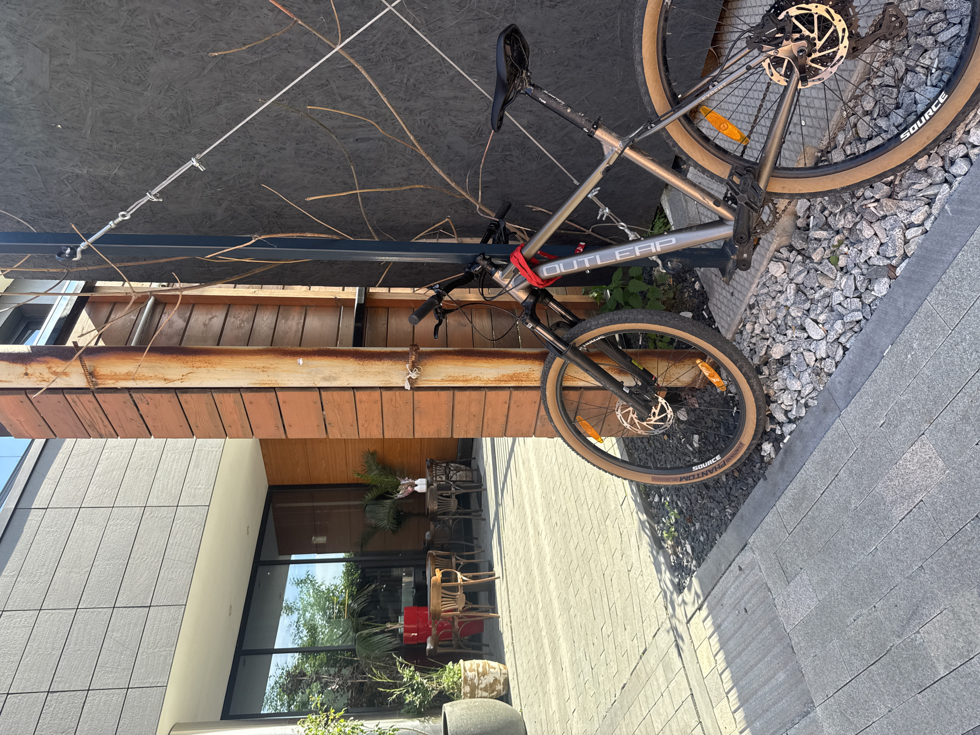 A silver mountain bike is parked outdoors, propped up against a dark wooden structure and a black wall. The bike is secured with a red strap around the frame. Its tires have tan sidewalls, and the rear wheel features a disc brake. The bike's frame has "OUTLEAP" written on it in large letters.
The scene appears to be an outdoor seating area, possibly a cafe or restaurant, with several chairs and tables visible in the background, some made of wood and others of a darker material. There are also some potted plants, including a palm tree. The ground is paved with grey rectangular stones.
The lighting suggests it is daytime, with sunlight casting shadows. The overall mood is casual and relaxed, indicating a pleasant outdoor environment. The image captures a moment of stillness, with the bike seemingly waiting to be ridden. The backdrop includes a modern building with gray tiled walls and large windows reflecting the sky and surrounding greenery. The context indicates the location is Yekaterinburg, Russia.