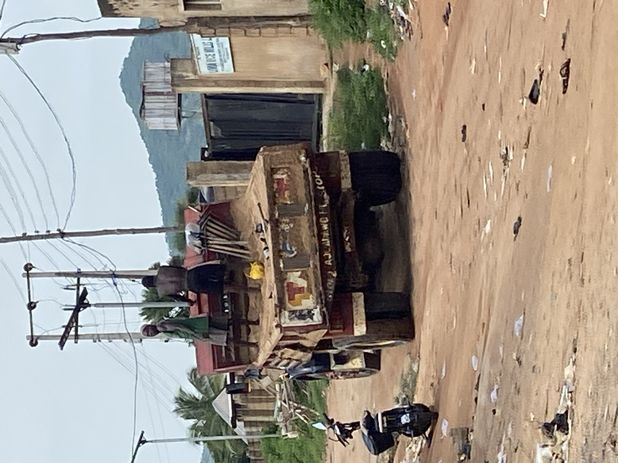 The image depicts a street scene in Kuson, Nigeria. A large, red, flatbed truck is the central subject, loaded with sand. Two individuals, a man and a woman, stand on the back of the truck. The man, shirtless, wears dark shorts. The woman is dressed in a green garment and a pink headwrap. A yellow bucket is visible on the sand. The truck has visible signage and markings. In the foreground, a black motorcycle is parked on the dusty ground. The environment appears to be an outdoor, unpaved street with debris scattered on the ground. A building with a sign reading "LIMAA RICE MILLS LTD." is visible to the right, and a green hill or mountain is in the background. Overhead, power lines and utility poles are present. The sky is overcast.