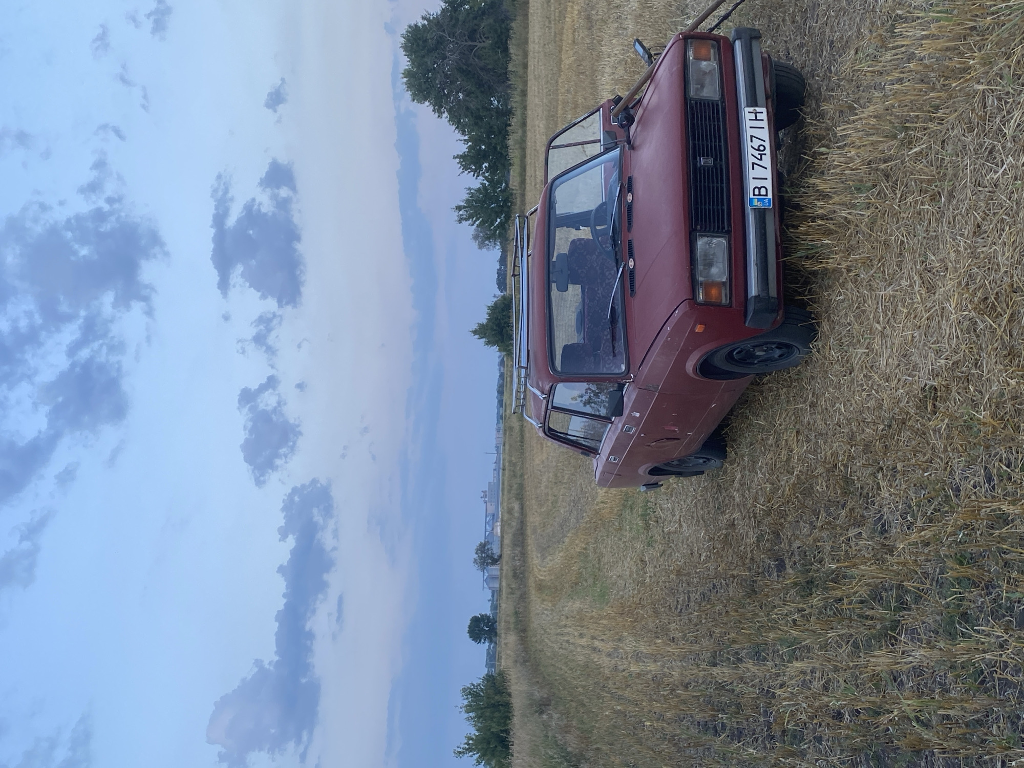 A red car is parked in a harvested wheat field under a cloudy sky. The car, a sedan, has its driver's side door open and a roof rack is attached to its roof. A dirt track leads into the field from the foreground. In the distance, several buildings and trees are visible. The license plate of the car reads "BI 7467 IH".