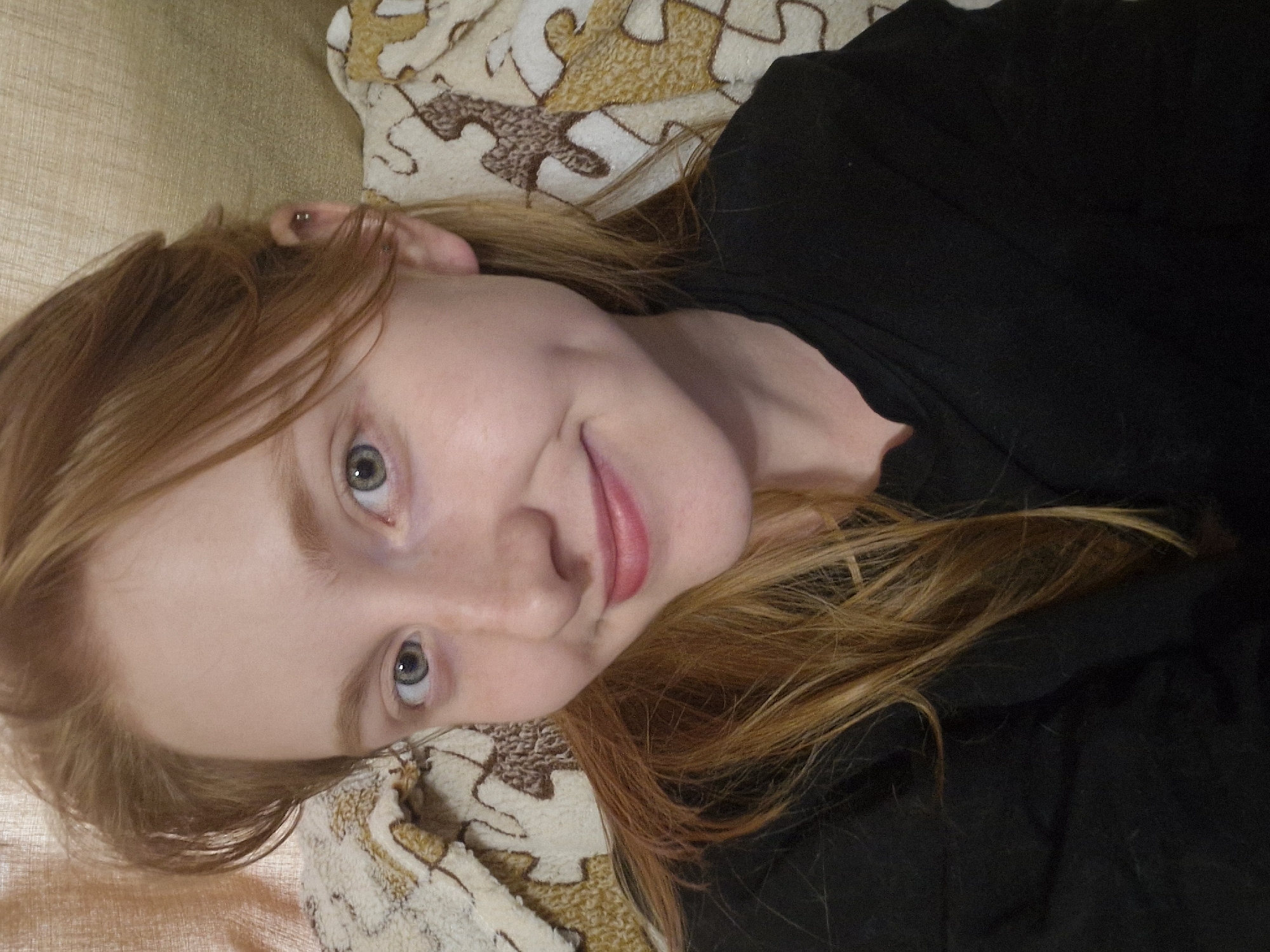 A young Caucasian female with red hair and fair skin is depicted in a close-up, selfie-style portrait. She has light blue eyes and a small dimple on her left cheek. She is wearing a black top and is leaning against a textured surface, possibly a blanket or cushion with a puzzle piece pattern in beige and brown. The background is a muted, textured wall in a warm, neutral tone. No specific location in Yekaterinburg, Russia, can be identified from the visual information provided in the image.