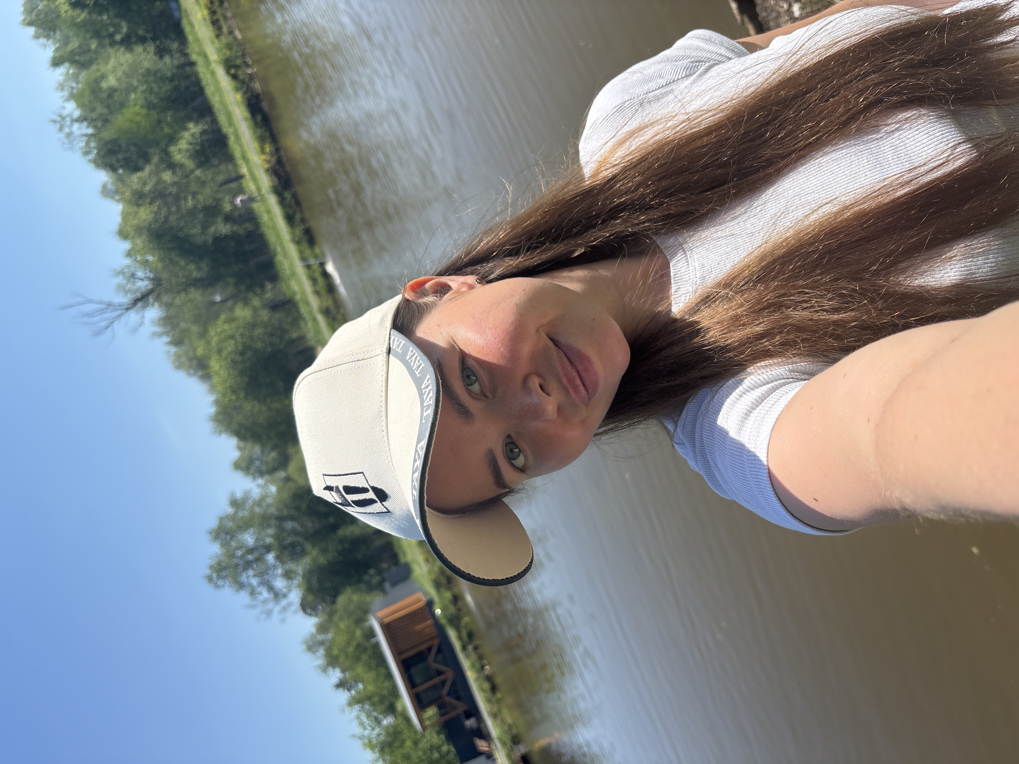 A woman wearing a light-colored baseball cap and a white ribbed shirt is taking a selfie outdoors. She has long brown hair and is standing near a body of water. Trees and a small structure are visible in the background. The scene appears to be set in Malyi Bukor, Russia, based on the request.