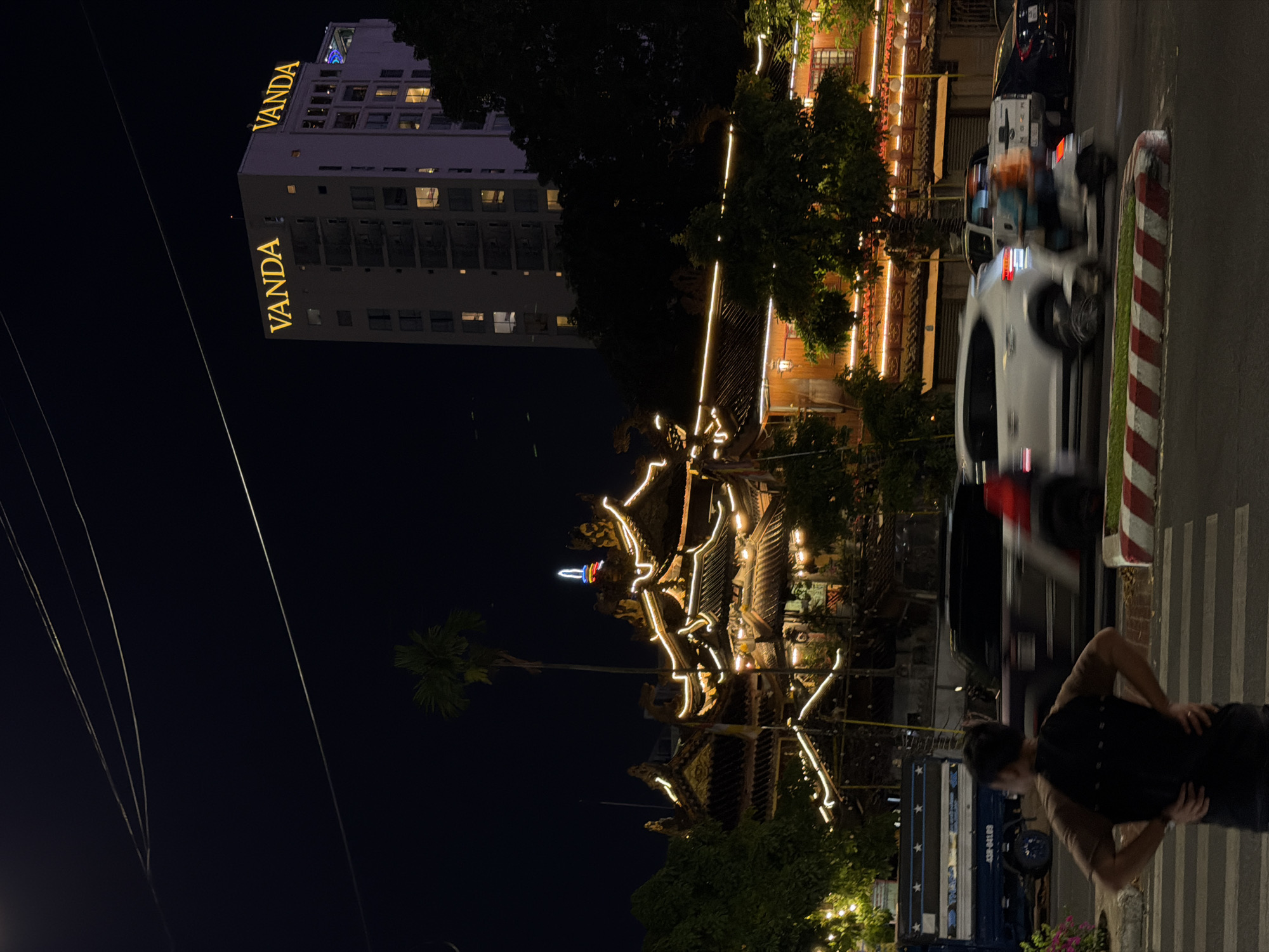 The image depicts a night scene in the city of Đà Nẵng, Vietnam. In the foreground, a man stands facing away from the camera with his hands on his hips, positioned at a crosswalk with red and white striped bollards. The street is illuminated by streetlights and the warm glow of lights from nearby buildings. Motion blur indicates vehicles, including a white SUV and a motorcycle with two riders, are in motion on the road. To the left, a blue truck with white stars and lettering is parked. In the midground, a traditional-style building with ornate, tiered roofs is decorated with numerous strings of yellow lights. A tall palm tree stands to the left of this building. Behind this structure, a modern, multi-story white building with illuminated windows is visible, displaying the prominent yellow signage "VANDA" at its top. The sky is dark, with thin power lines stretching across it. Trees are interspersed throughout the scene, with some lit with string lights.