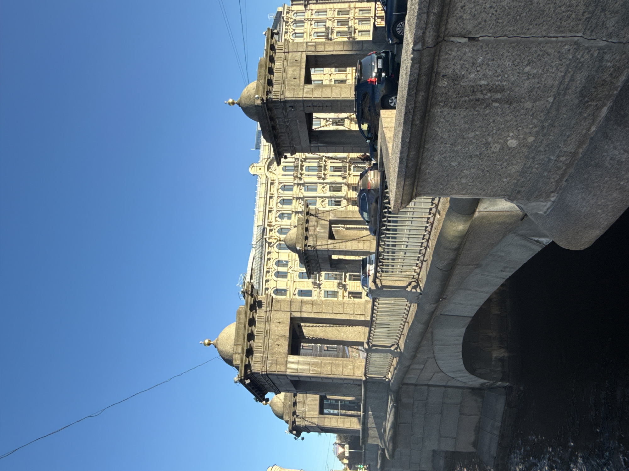The image displays an urban landscape featuring an ornate stone bridge, a grand building, and a clear blue sky, set in Saint Petersburg, Russia. The scene is captured during daylight hours under bright, clear weather.
The primary subject is a sturdy bridge constructed from large, grey, textured stone blocks. It features prominent, square-shaped pillars or pylons along its edge, each topped with a distinctive, light-colored dome that culminates in a small, golden finial. Decorative chains are suspended between these pillars, forming part of the bridge's railing. Several dark-colored cars are visible on the bridge or the road running alongside the building on the far bank.
Beyond the bridge, a large, multi-story building with classical architectural elements stands prominently. Its facade is a light yellow or beige, adorned with numerous arched windows and intricate detailing such as pilasters and cornices. The building's ornate design is characteristic of historical European architecture.
Beneath the bridge, a dark body of water flows. The sky above is a vibrant, cloudless blue, indicating a sunny day. A thin, dark wire or cable is faintly visible traversing the upper portion of the sky. No specific activity or event is depicted beyond the general presence of cars and static city structures. There is no clearly discernible text within the image.