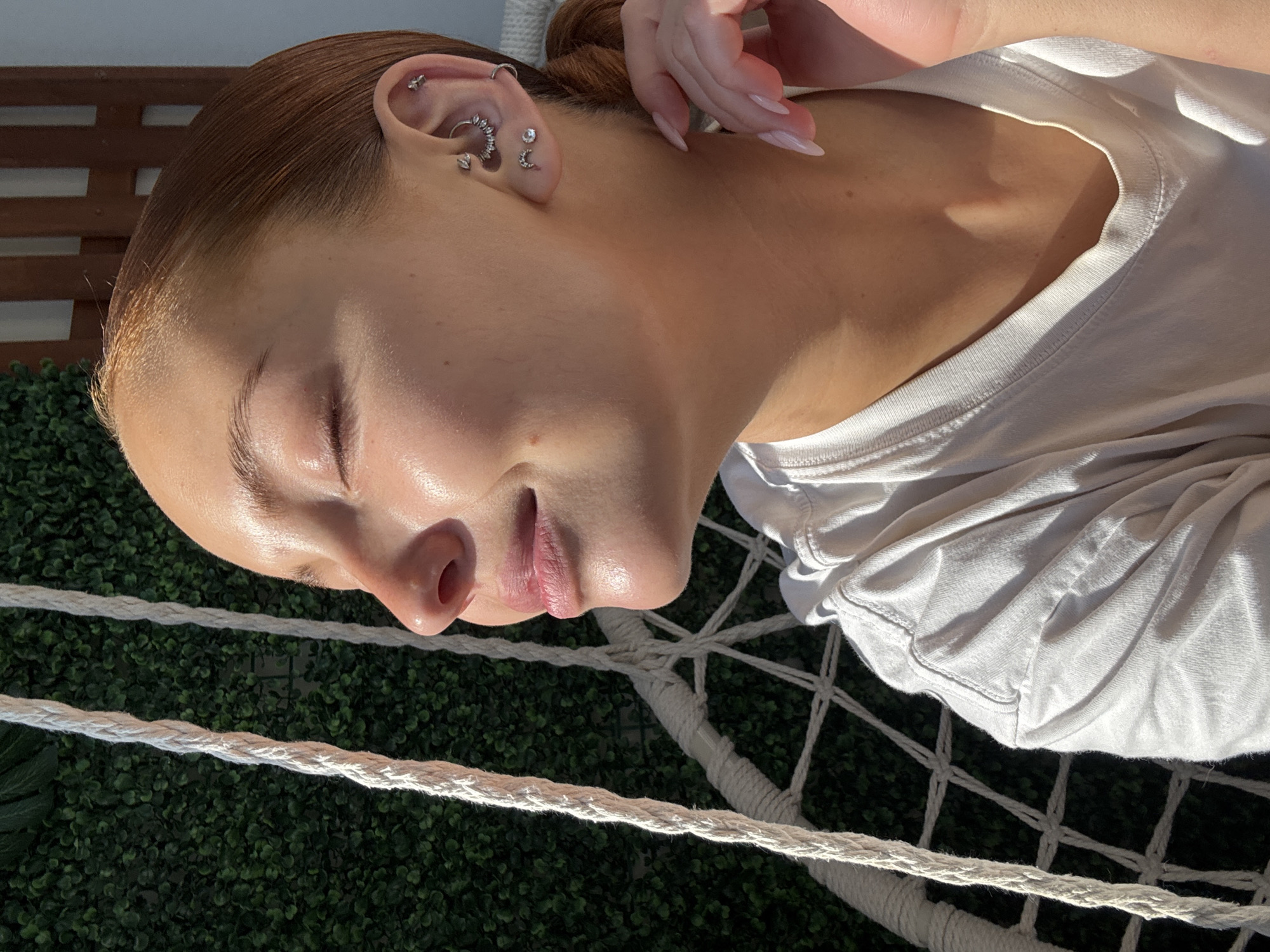 A medium shot depicts a young woman with fair skin, closed eyes, and multiple ear piercings. Her hair is light brown and pulled back. She is wearing a white t-shirt. The background consists of green foliage and the white ropes of a hammock. The photo appears to be taken outdoors in natural light. There is no indication that the location is Chaikovsky, Russia.