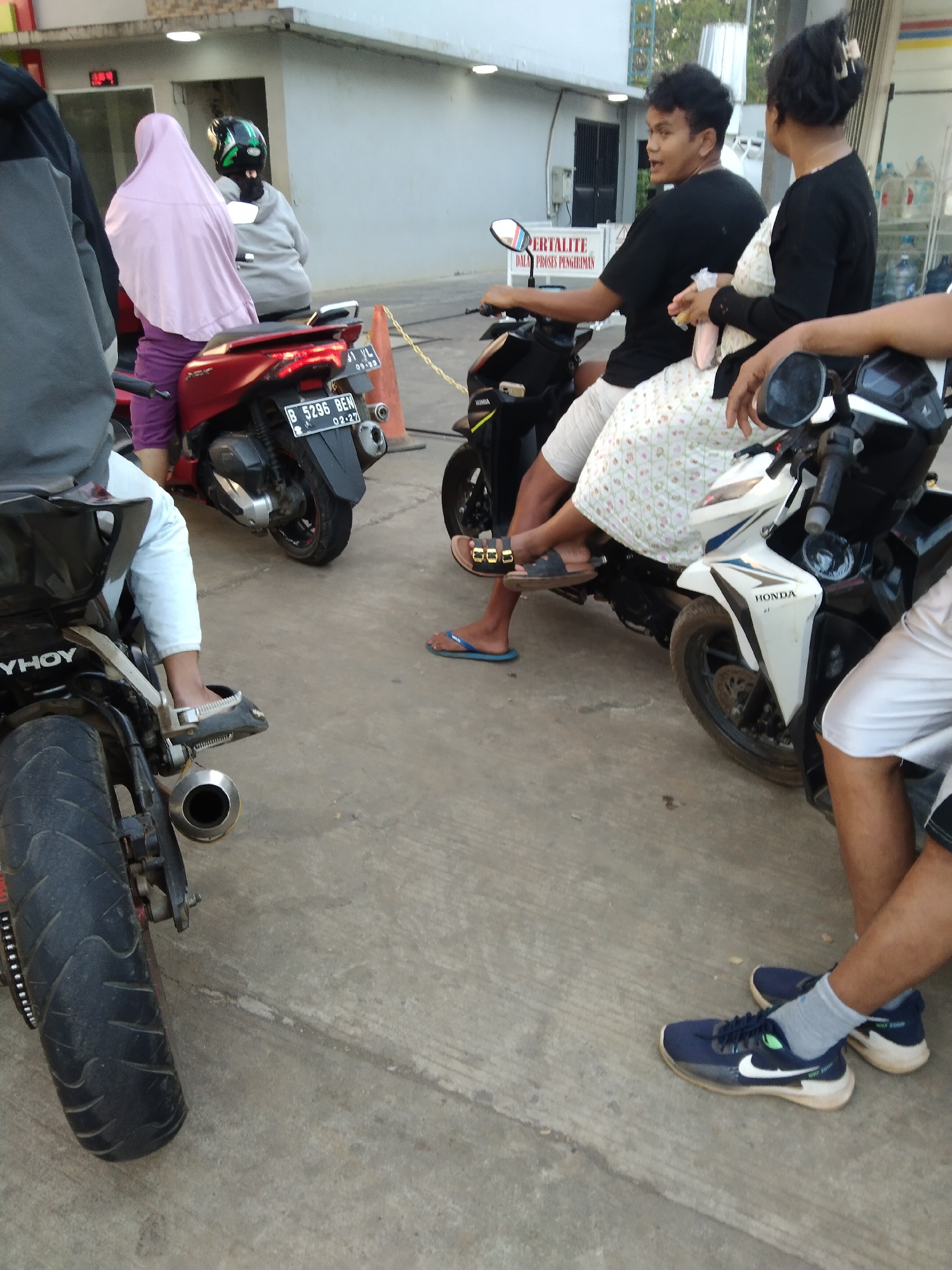 The image captures a scene at an outdoor area, likely a gas station or roadside shop in Kembang, Indonesia. Several individuals are present, some on or near motorcycles. In the foreground, a person wearing blue Nike sneakers and light-colored shorts is seated. To the left, a motorcycle is partially visible with its rear wheel and exhaust. Further back, a woman in a pink hijab and purple pants is seated on a red motorcycle. Next to her, another person is on a motorcycle wearing a gray hoodie and a green helmet. In the center, a man in a black t-shirt and light-colored pants is on a motorcycle, and a woman beside him is wearing a white dress with a floral pattern. Another motorcycle, predominantly white and black with "HONDA" branding, is in the mid-ground. The ground is paved with concrete, and a yellow traffic cone is visible. In the background, a building with windows and what appears to be a signage with Indonesian text can be seen. The lighting suggests it is daytime.