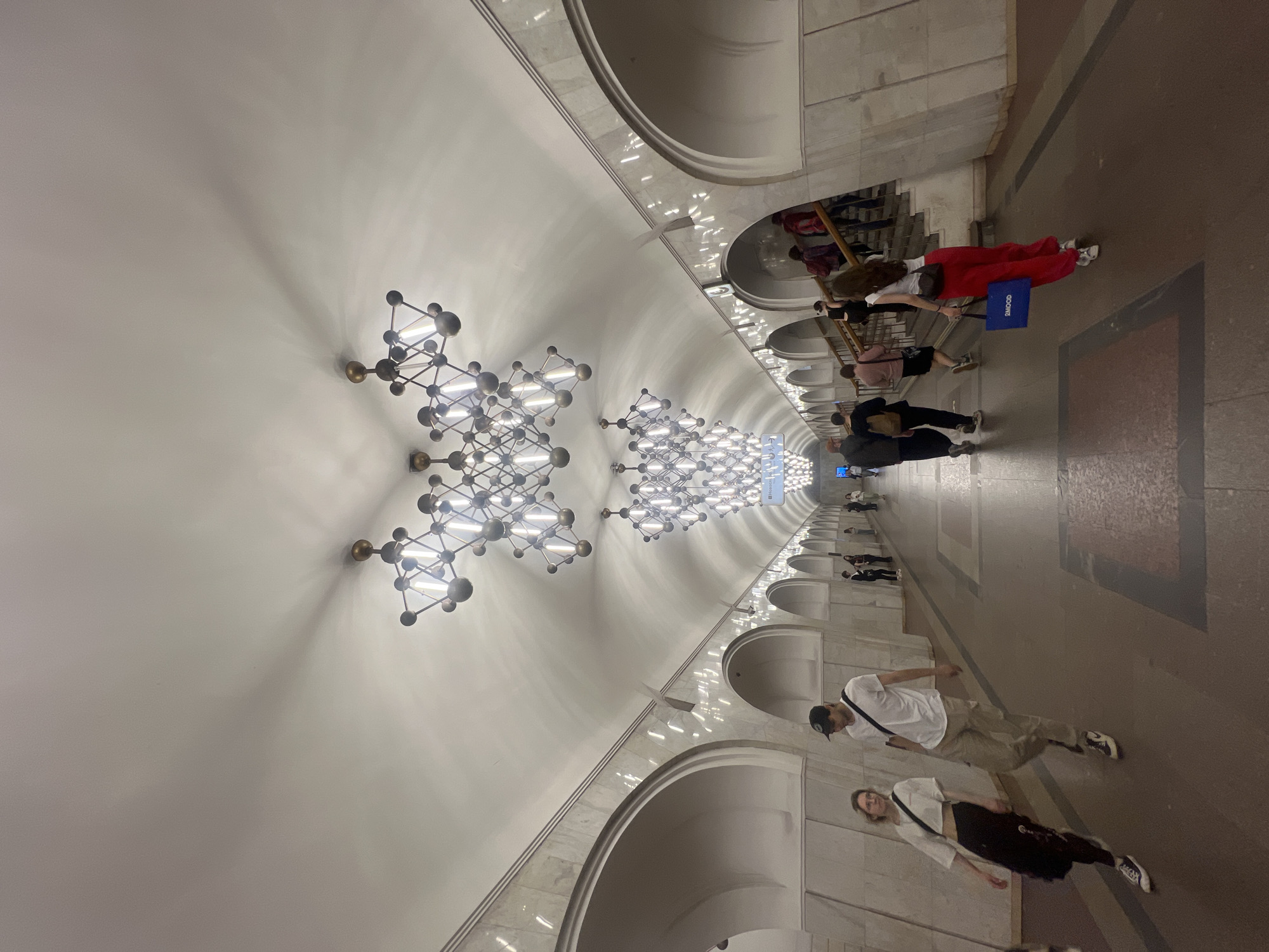 A photo depicts a station in the Unknown, Unknown area. The image, presented in a tilted perspective, showcases the station's interior, characterized by arched ceilings and ornate light fixtures hanging overhead. Multiple individuals are observed walking along the station platform, suggesting pedestrian traffic. The platform's surface appears to be a mix of textures and materials. The lighting conditions illuminate the scene, highlighting the architectural features and the people within it.