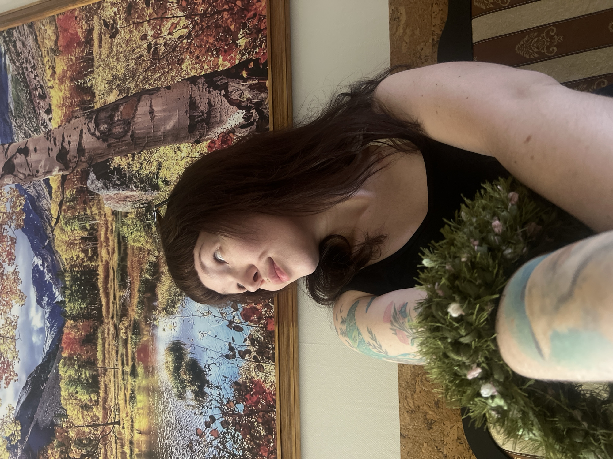 The image shows a young woman with long, dark brown hair and bangs, wearing a black tank top. She has tattoos on her arms, which appear to be floral designs in blue, green, and pink. She is holding a green wreath with small pink and white flowers. The woman is looking up and to her right, with a slight smile on her face. She is sitting on a bench with a striped cushion.
In the background, there is a large framed print or painting of a landscape. The artwork depicts a body of water in the foreground, with trees and mountains in the background. The trees in the foreground appear to be birch trees, one of which is prominent in the right side of the image. The landscape has a warm, autumnal color palette, with vibrant reds, oranges, and yellows in the foliage, contrasting with the blue of the water and the grey of the mountains.
The overall scene is indoors, likely in a residential setting. The bench and the wallpaper on the wall suggest a domestic environment. The lighting is warm and somewhat diffused, contributing to a cozy atmosphere. The image is taken from a medium-low angle, focusing on the woman and the artwork.
Regarding the location in the city of Chaikovsky, Russia, there is no specific information in the image that definitively places it there. However, the presence of a large landscape painting featuring birch trees and a body of water might evoke a sense of the Russian natural landscape, which is common in art and decor in Russia. Without further context or identifying landmarks, it is impossible to confirm the specific location within Chaikovsky.