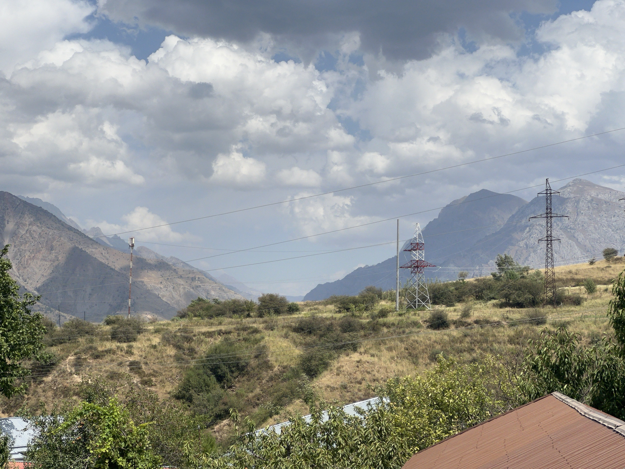 This is a landscape photograph taken in Yusuphana, Uzbekistan, featuring a natural environment with man-made structures. The image is dominated by a cloudy sky and mountainous terrain. In the foreground, there is dense green foliage and a portion of a reddish-brown corrugated roof. The midground shows a dry, grassy hillside dotted with scattered bushes and trees. Two tall power pylons are visible; one is a lattice tower with red and white accents, and the other is a darker, more utilitarian metal structure. A communications tower with red and white bands stands on the hillside further in the distance. The background consists of rugged, sparsely vegetated mountains under a dramatic sky filled with white and gray clouds. The overall impression is of a rural or semi-rural area with a mix of natural beauty and industrial infrastructure.