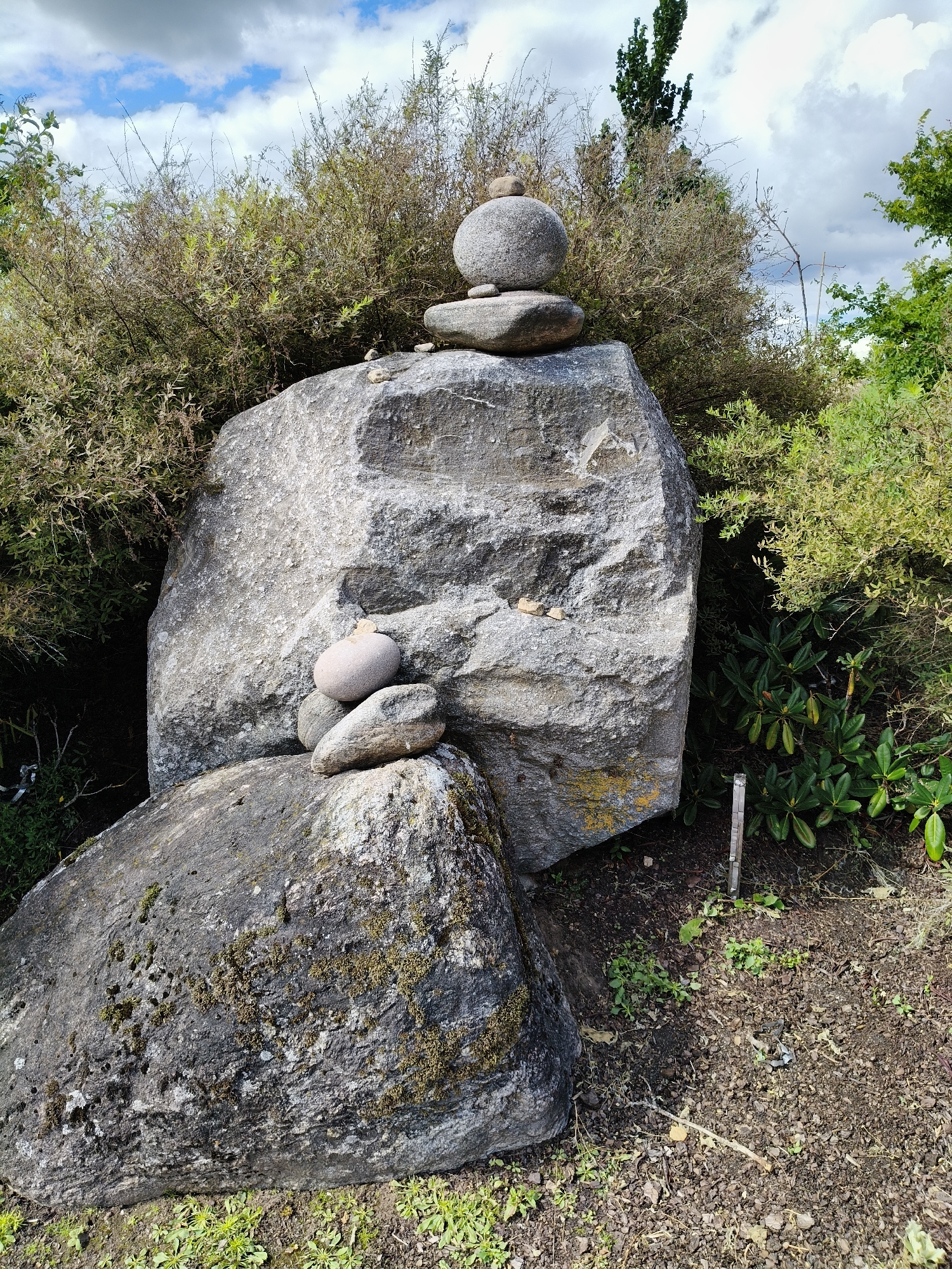 The image depicts an outdoor scene with a focus on a stack of stones in front of dense vegetation. Two large grey boulders form the base of the composition. The boulder on the right is significantly larger and has a roughly textured surface with some yellowish-green lichen. On top of this boulder, a stack of smaller, smoother grey stones is carefully balanced. The topmost stone is round, followed by a slightly flatter, oval-shaped stone, and then another flatter, irregular-shaped stone.
Behind the boulders, a thicket of dry, thorny bushes with sparse green leaves dominates the background. To the right of the bushes, a taller, dark green tree is visible. The ground in front of the boulders is covered with dry soil, small pebbles, and some sparse green vegetation. A thin wooden stake with a small, reflective metal plate is partially visible in the soil to the right of the large boulder. The sky above is partly cloudy with patches of blue sky visible. The scene appears to be in a natural or semi-natural setting. The image is shot from a slightly low angle, looking up towards the stacked stones and vegetation.