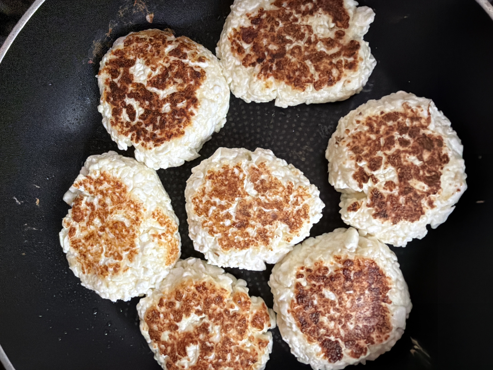 A top-down, close-up shot shows seven round, flattened foods, possibly fritters or patties, cooking in a dark non-stick frying pan. The foods are off-white with irregular, golden-brown to dark brown toasted patches on their surfaces. The pan has a matte black finish and a textured surface with a slight sheen, indicating it is heated. The lighting is natural and comes from above. There is no visible indication of a specific location in Khabarovsk, Russia.