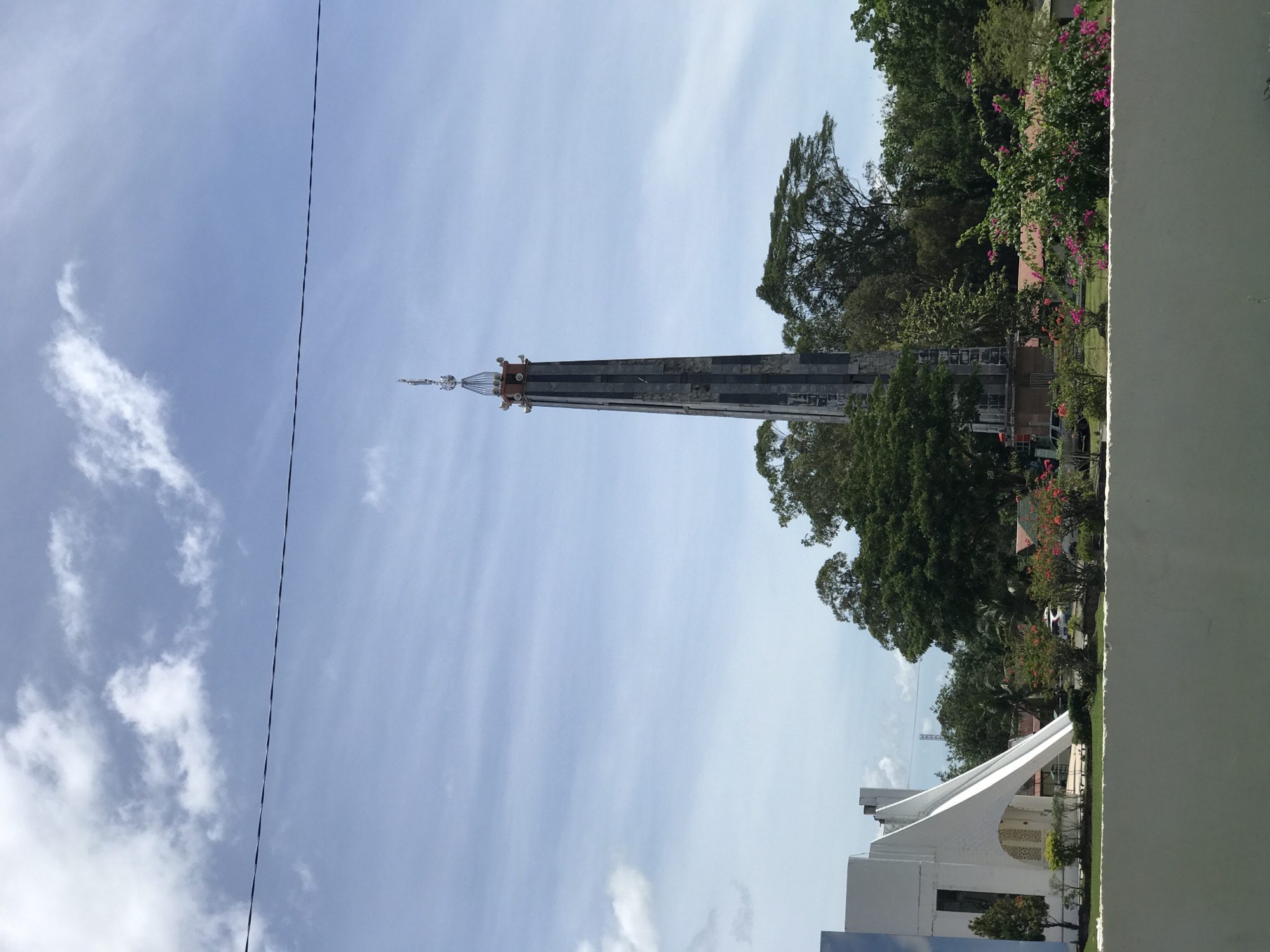 The image shows the Baiturrahman Jantho Grand Mosque in Jantho, Aceh Province, Indonesia. The mosque's minaret is the primary focus, a tall, slender structure with horizontal banding and a decorative finial at the top. The surrounding environment includes green trees, flowering plants, and a partially visible white building with a distinctive arched roof. The sky is blue with scattered clouds. A power line is visible in the upper-left quadrant.