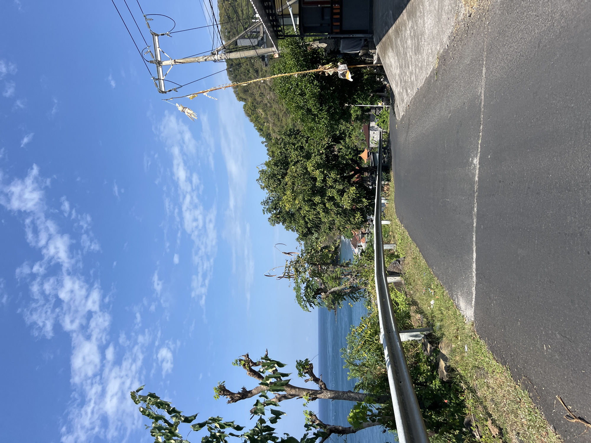 A paved road curves uphill, bordered by a metal guardrail on the left and a concrete walkway on the right. The road faces a lush green hillside dotted with buildings and trees. The ocean is visible in the distance to the left, with a clear blue sky and scattered clouds above. A utility pole with wires and festive decorations stands on the right side of the road, near a building. The scene appears to be in Abang, Indonesia, a coastal region known for its natural beauty and local culture.