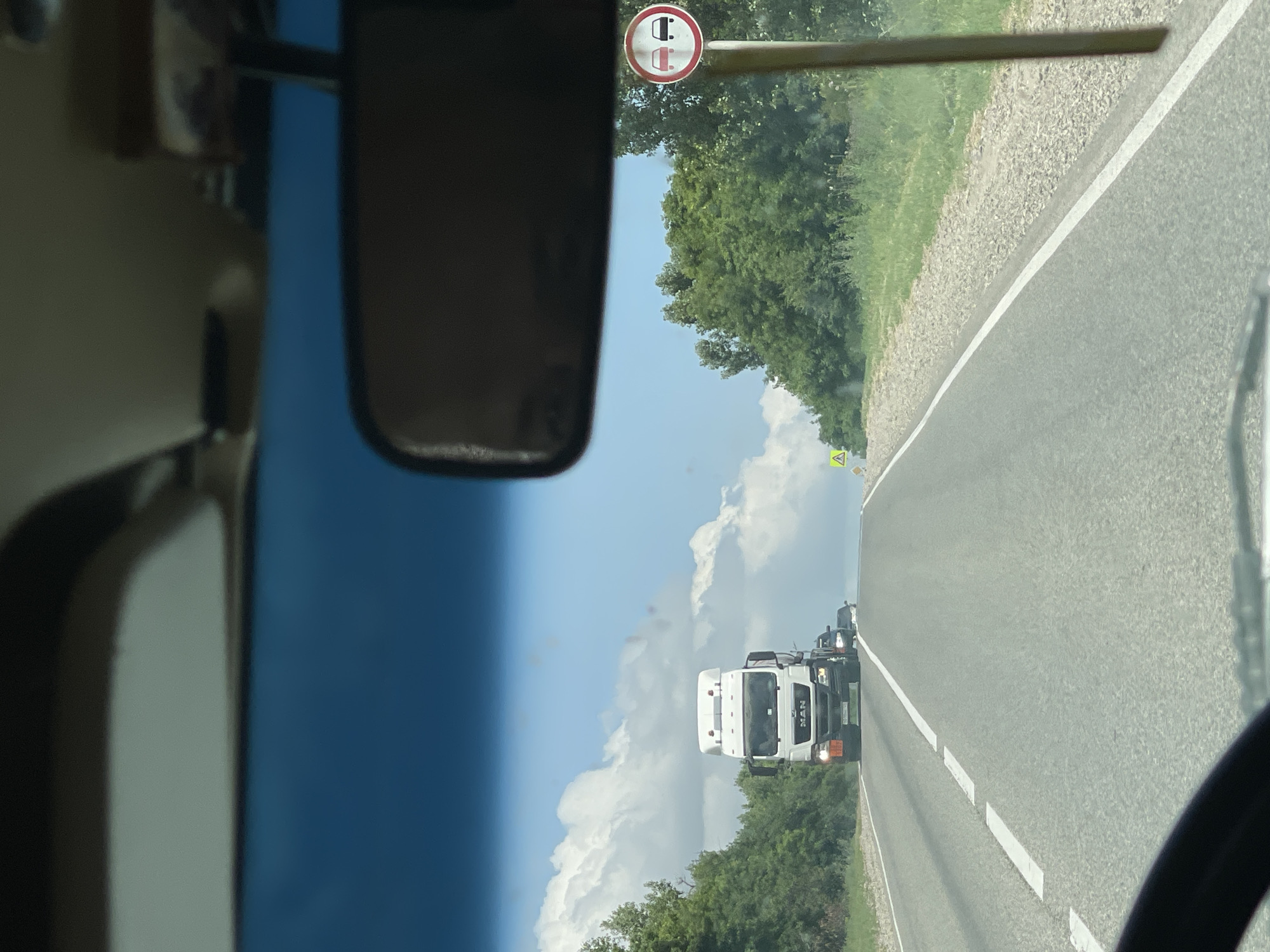 The image is a first-person perspective from inside a vehicle, looking forward on a paved two-lane road under a cloudy blue sky. A large white MAN truck is driving ahead in the right lane, followed by at least one dark-colored passenger vehicle. To the left of the road, there is a line of green trees and vegetation. To the right of the road, there is a signpost with a circular traffic sign indicating "No Overtaking" for all vehicles, depicted by two cars, one red and one black, within a red circle and a red diagonal line. Further ahead on the right side, a yellow triangular warning sign with a black symbol is visible. The road appears to be in a rural or suburban area. The location is identified as Aleksandrovskoye, Russia.