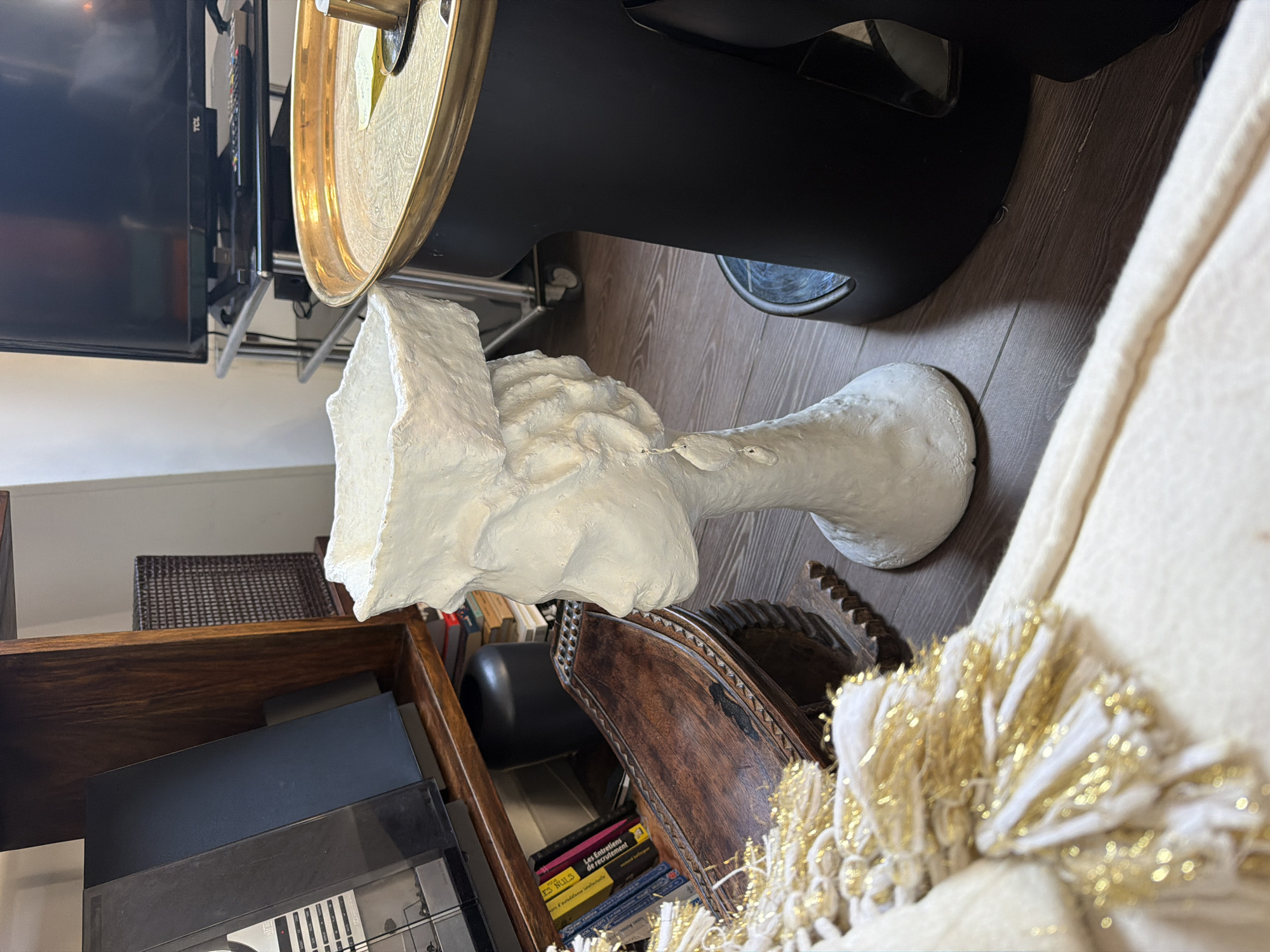 The image captures an indoor living space or study, showing a collection of decorative and functional items. The central subject is a rough-textured, white bust, resembling an ancient Egyptian Nefertiti bust, positioned on a dark wooden shelf or cabinet.
To the left and slightly behind the bust, a flat-screen television with "TCL" branding is visible, mounted on a stand. Adjacent to the TV, a prominent golden, ornate tray rests on what appears to be a dark piece of furniture or an audio component. The floor in this area is covered with wooden laminate planks.
On the right side of the image, a cream-colored piece of furniture, likely a couch or armchair, features a decorative throw blanket with an elaborate fringe of white and shimmering gold threads or tinsel. Below the main shelf holding the bust, another dark wooden shelf contains various books and other items. Among the visible books, one spine clearly displays the French title "Les Entretiens de recrutement," and another partially shows "ES NULS," which aligns with the provided location context of Paris, France.
No specific activity or event is taking place; the scene presents a static interior arrangement. The lighting suggests it is daytime. The overall impression is that of a comfortably furnished room with a mix of classic and modern aesthetics.