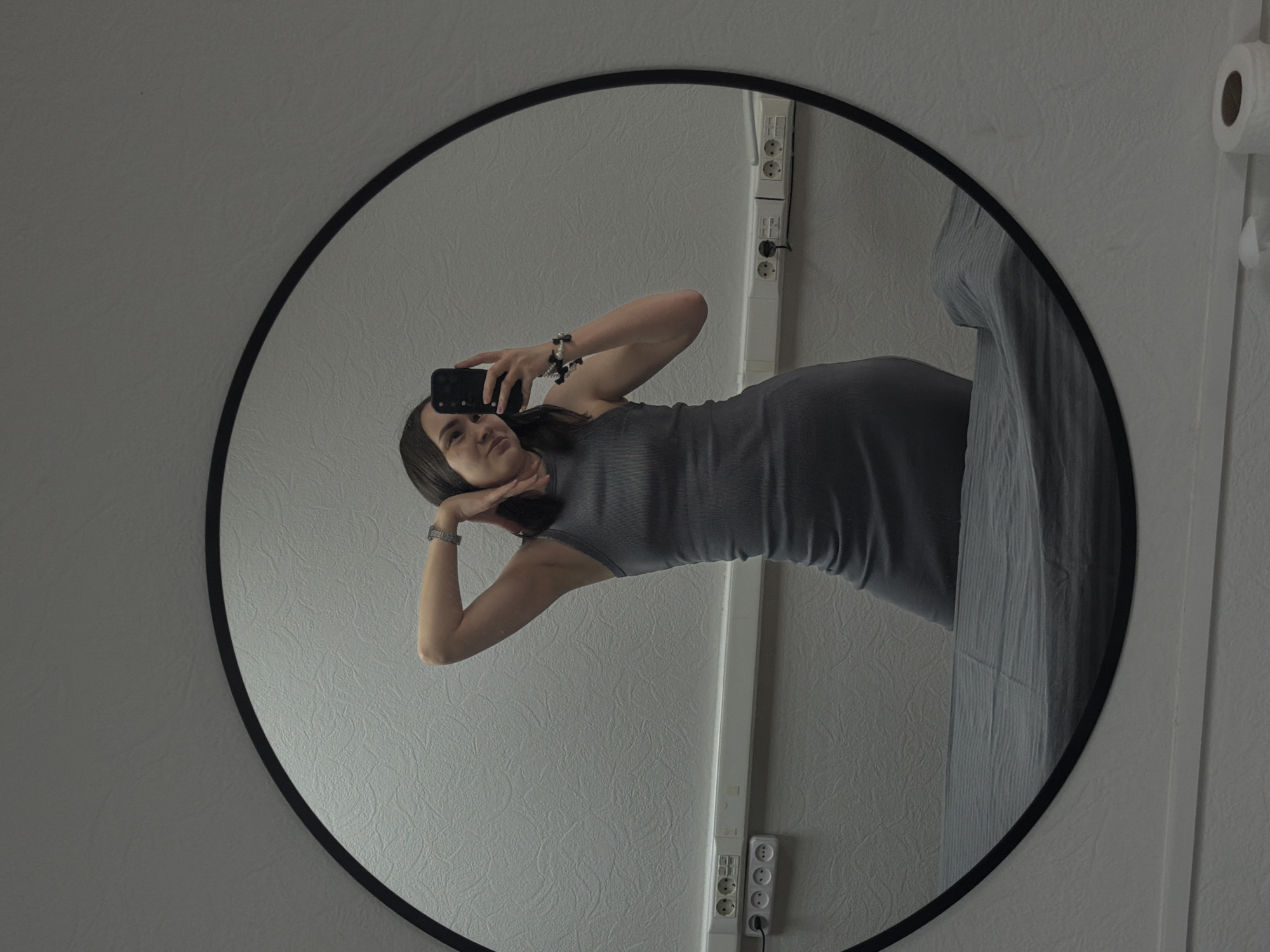 The image is a selfie of a woman in a gray dress taken in a round mirror. The reflection shows the woman holding a phone, posing with her hands near her face. The background reveals a white textured wall with electrical outlets and a roll of toilet paper. It appears to be a bathroom setting. The image metadata indicates the location as Chaikovsky, Russia.