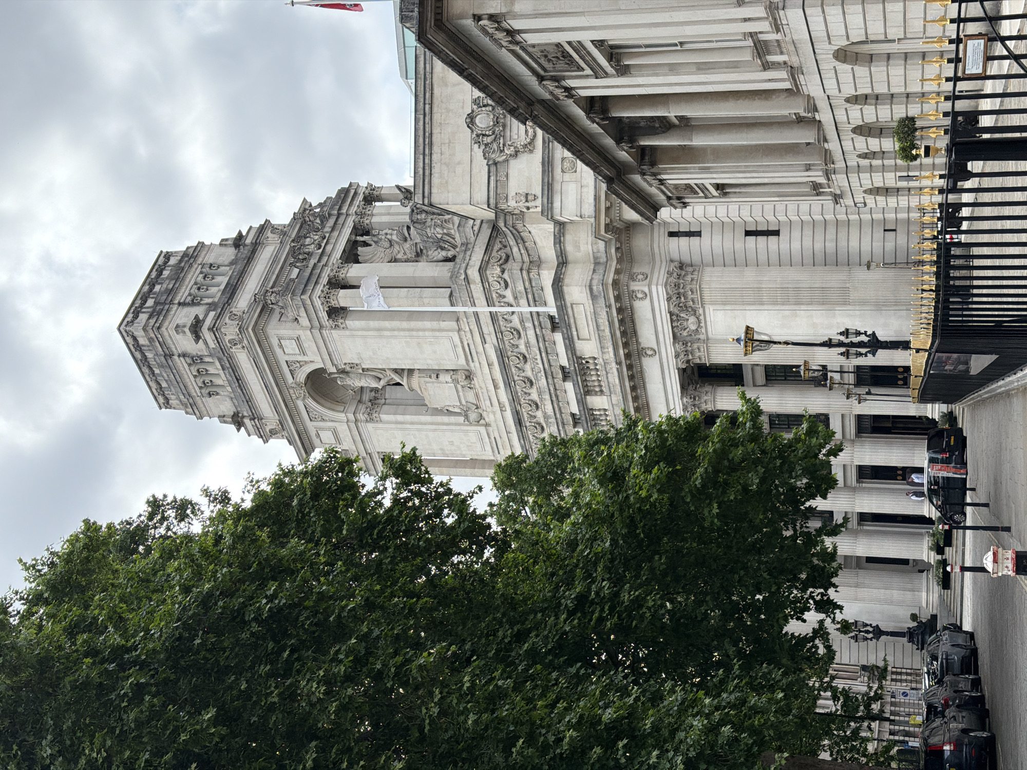 This image depicts the Port of London Authority building, also known as Ten Trinity Square, located in the City of London, United Kingdom.
The scene is dominated by a grand, multi-story Neoclassical building constructed from light-colored, weathered stone. Its most prominent feature is an elaborate central tower, characterized by multiple tiers, large columns, arched openings, and numerous sculpted figures. A white flag with a dark logo is visible flying from a flagpole near the tower's mid-section, and a red flag with a white cross (St George's Cross) is partially visible on the building's roofline to the right. The building's lower facades feature prominent columns, ornate friezes, and detailed architectural ornamentation.
A large, dense green deciduous tree occupies the entire left side of the frame, partially obscuring the building's left wing and some of its ground-level features. Its foliage appears healthy and full.
At street level, a paved road runs across the bottom of the image. Several dark-colored vehicles, primarily SUVs and sedans, are parked parallel to the curb on the left side. Further down the street on the right, a black minivan-style vehicle with an orange sticker on its side is visible. Ornate black street lamps with multiple light fixtures are positioned along the pavement on the left. Two individuals, appearing to be staff in light-colored uniforms, stand near an entrance on the left side of the building, partially visible through the trees.
On the right side of the scene, a black metal fence with decorative golden spear-top finials separates the pavement from the road and the building. A small informational plaque is attached to this fence.
The sky is uniformly overcast and grey, with visible cloud formations, indicating diffused lighting conditions across the scene.