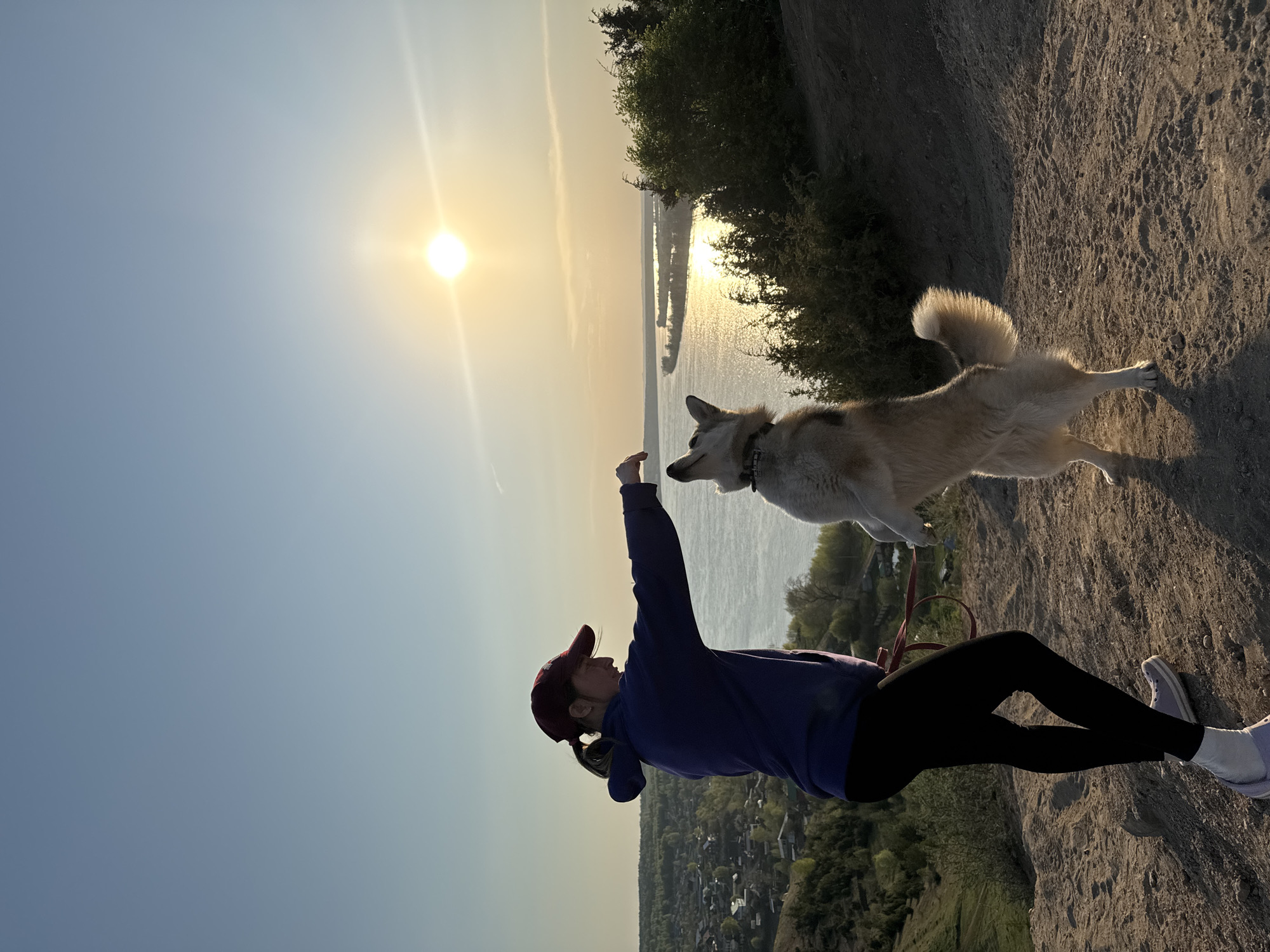 A woman and a dog are on a rocky hill, likely in the city of Olkhovka, Russia. The woman is wearing a dark blue sweatshirt, black leggings, white shoes, and a maroon baseball cap. She is raising her right arm, presumably to interact with the dog. The dog is a medium-sized, light-colored breed. The setting appears to be a hill overlooking a body of water, possibly a river or lake, with the sun setting on the horizon. The environment suggests a park-like or natural area within or near a city.