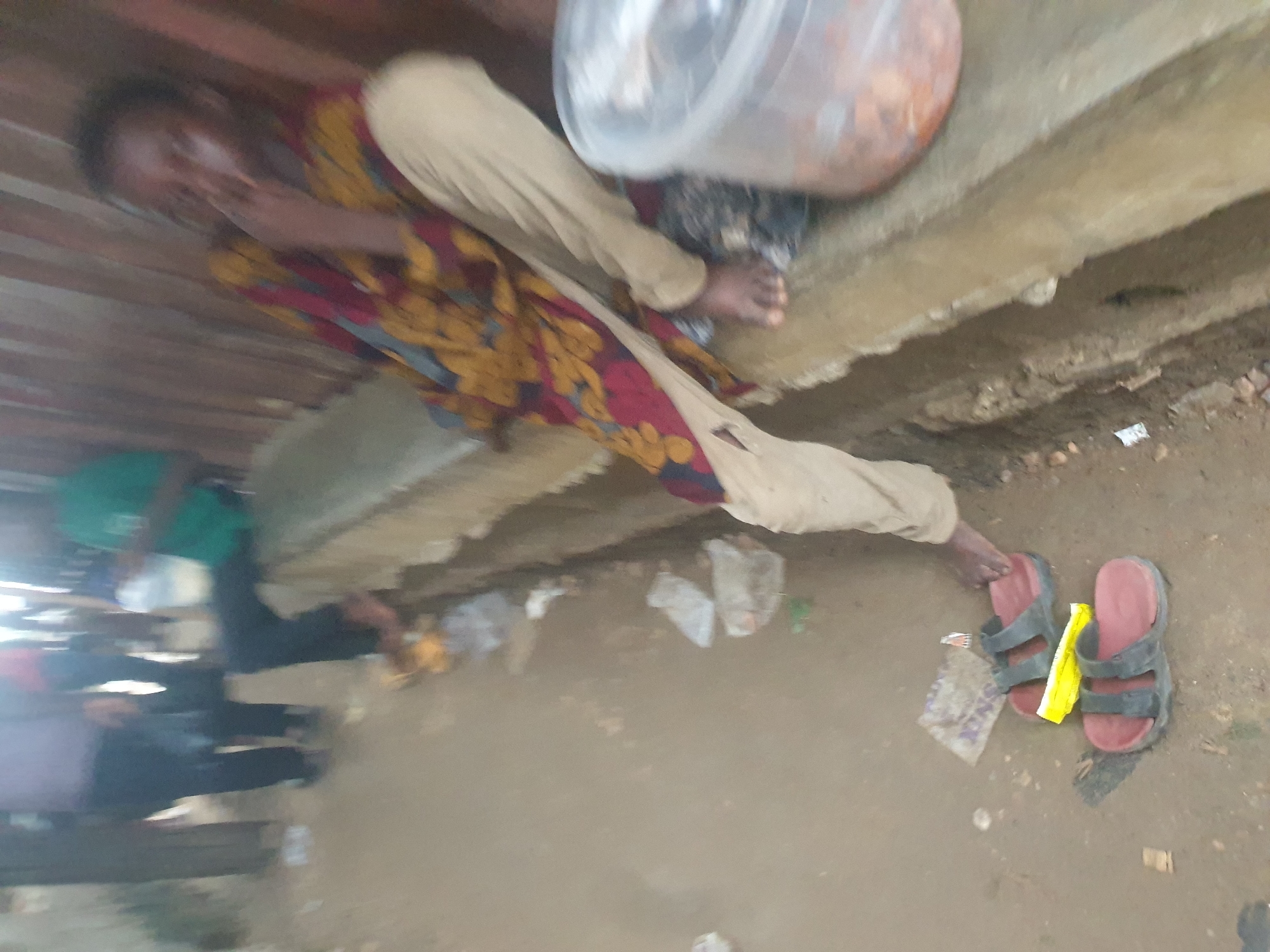 The image shows a person reclining on a concrete ledge in Adankolo, Nigeria. The individual wears a patterned top and light-colored pants. A clear plastic container sits on the ledge near them. Their bare foot is visible, extending toward a pair of red and black sandals on the ground. Debris, including plastic bags and wrappers, litters the dirt ground. The background includes corrugated metal. Other people are visible in the background.