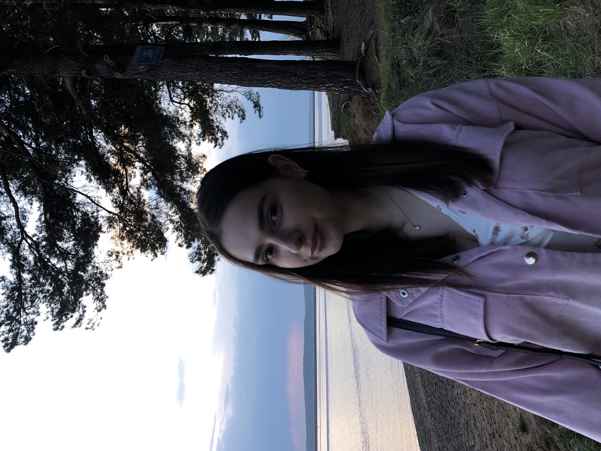 A young woman is photographed outdoors in what appears to be the city of Chaikovsky, Russia. She is wearing a light-colored floral top and a pink jacket, and is positioned on a bank or cliff, with a body of water visible behind her. A stand of pine trees is to her left, some bearing a sign, and green grass is to her right. The sky is partly cloudy.