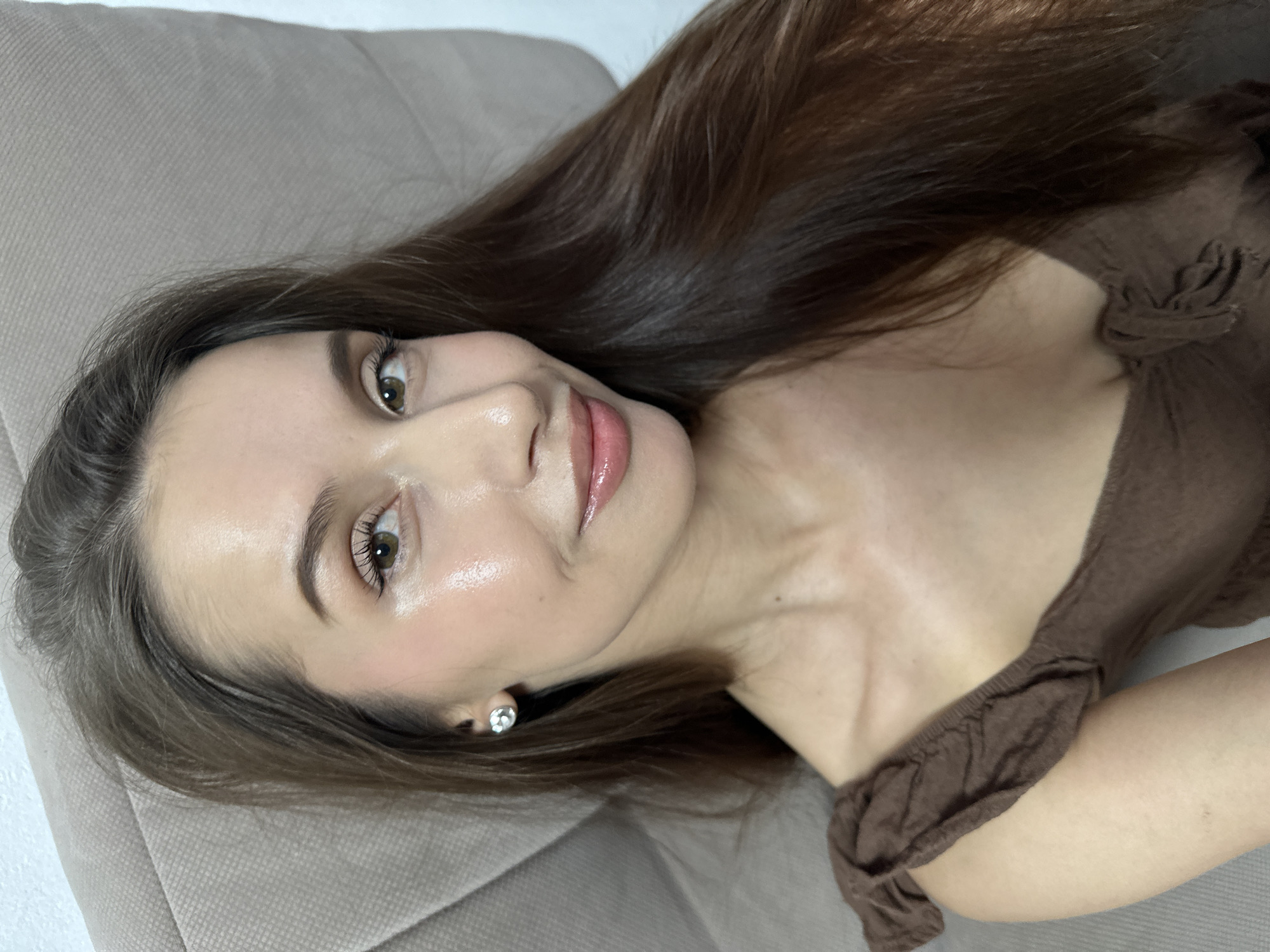 A close-up portrait shows a young adult woman with long, dark brown hair, lying or reclining on a light-colored upholstered surface, likely a sofa. Her head is tilted, and she looks directly at the camera with a subtle, serene smile and brown eyes. She has a natural makeup look, featuring glossy pink lips and defined eyelashes. A small, clear stud earring is visible in her left ear. She is wearing a brown, off-the-shoulder top, with some of her shoulder and arm visible in the frame. The lighting is soft and even, suggesting an indoor daytime setting. The background is simple and out of focus, emphasizing her features. The location context for this image is Yekaterinburg, Russia. No visible text is present.
