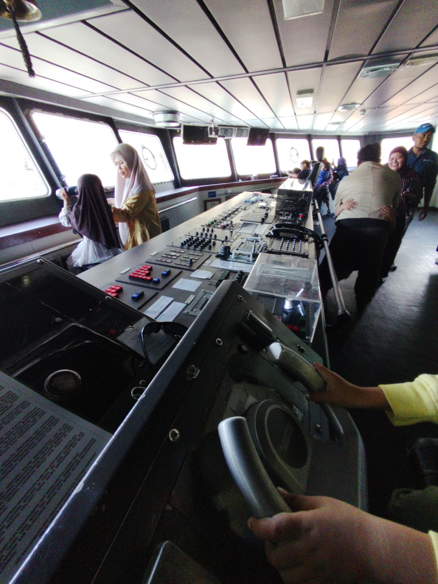 The image depicts the interior of a ship's bridge. Two women wearing headscarves are standing near a large window overlooking what is likely the ocean. One woman, dressed in a yellow hijab and yellow garment, is looking out the window. The other woman, wearing a dark purple hijab and white garment, is also looking out. Several control panels and equipment are visible in the foreground, suggesting the bridge is functional. In the background, other people are present, including a man in a blue uniform and hat. The lighting suggests it is daytime, with natural light coming from the windows. The environment appears to be a professional setting on a vessel.