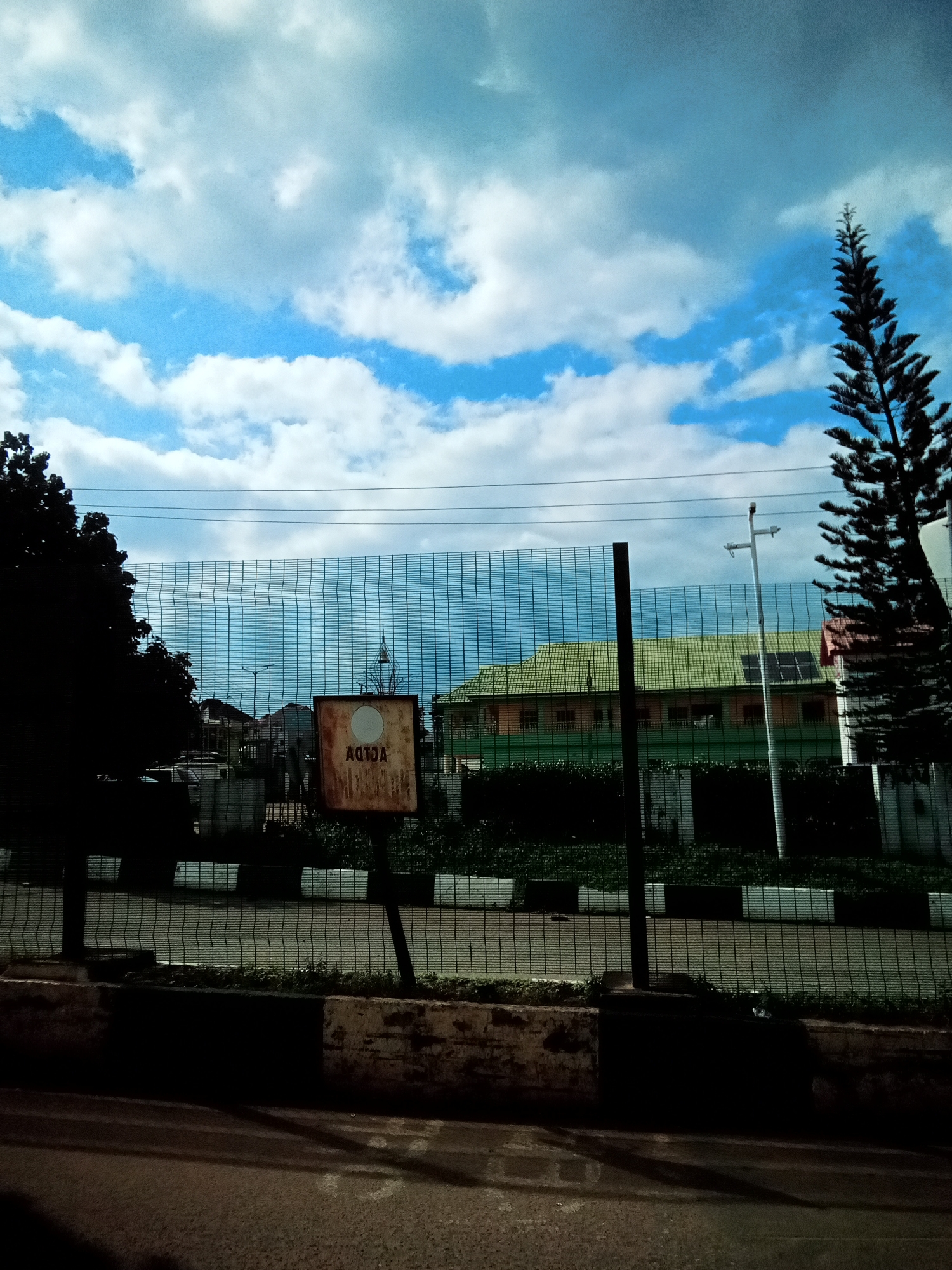 The image shows a view of a building with a green roof and solar panels, partially obscured by a metal fence. A dark silhouette of a tall evergreen tree is on the right side of the frame, and a tree with dark foliage is on the left. A rusty sign with the text "A010A" and a white circle is visible behind the fence. The scene appears to be in Nibo, Nigeria, as indicated by the sign, which is likely a street or building marker. The sky is partly cloudy with a mix of blue sky and white clouds. The foreground shows a dark road and a concrete barrier.
