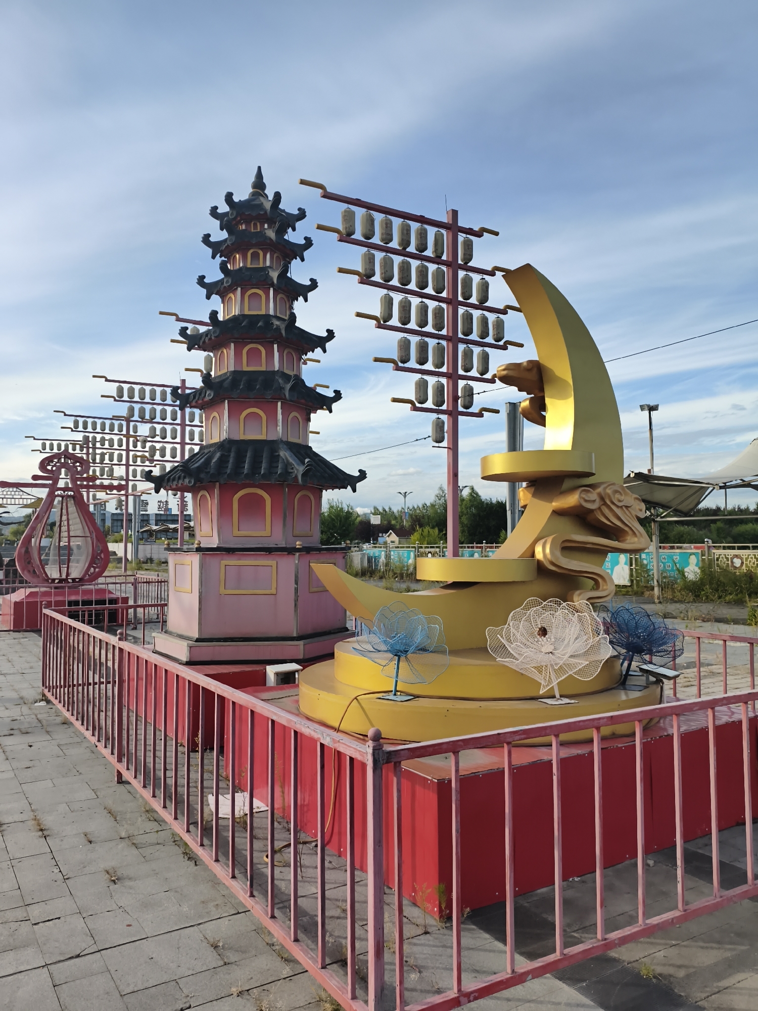 This image captures an outdoor scene featuring a decorative structure with East Asian influences, situated in 新城村 (Xīnchéng Cūn), China. The primary elements of the structure include a multi-tiered pagoda painted in pink and yellow with black roofs, and a large golden crescent moon with a sculpted figure, possibly a dragon or frog, perched on it. The pagoda is adorned with decorative windows and tiered roofs characteristic of traditional pagodas. To the left of the pagoda, a red, abstract lantern-like sculpture is visible. In front of these main structures, a yellow base is decorated with three wire-frame floral sculptures in blue, white, and blue. These elements are enclosed by a red metal fence. In the background, a series of horizontal poles are strung with numerous small, light-colored lanterns. The scene is set against a bright blue sky with scattered clouds, and the ground is paved with grey square tiles. The overall atmosphere suggests a public space, possibly a park or a decorative area within a village, illuminated by daylight.