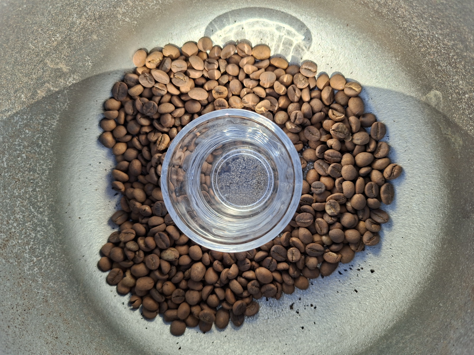 This is a top-down, close-up shot of roasted coffee beans arranged in a heart shape within a metallic, circular container. A clear glass, partially filled with what appears to be a small amount of dark liquid, sits at the center of the heart of coffee beans. The container has a textured, matte gray surface, and a shadow cast by the glass and beans falls across the right side of the frame, creating a play of light and dark. The image does not depict any identifiable locations within Oxentea, Moldova, or any individuals or actions.