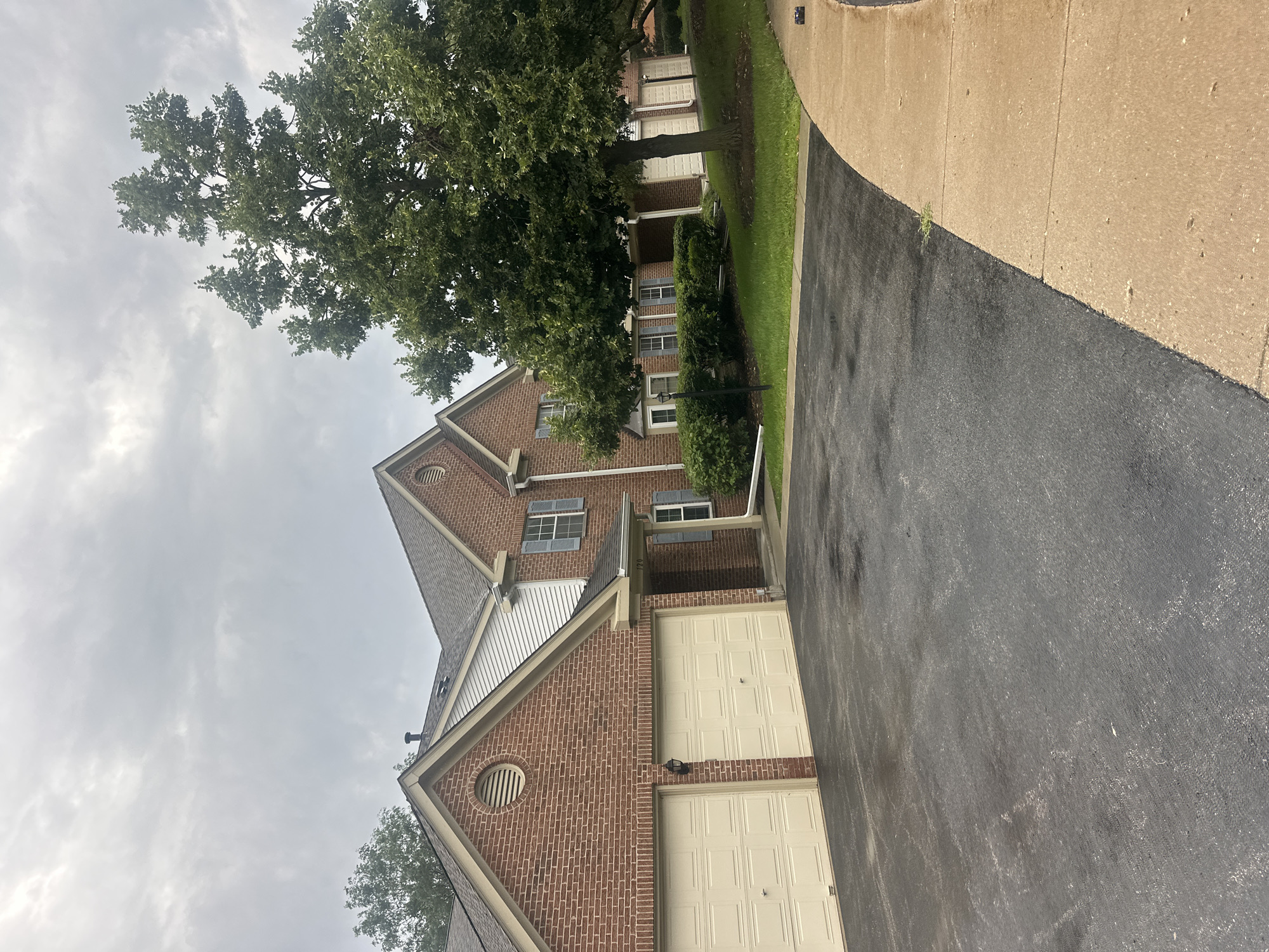 The image shows a row of brick townhouses in Schaumburg, Illinois, United States of America. The townhouses have attached garages with beige doors. There is a large tree in front of the townhouses, and the sky is overcast. The ground in front of the townhouses is paved with asphalt. There is a sidewalk to the right of the asphalt.