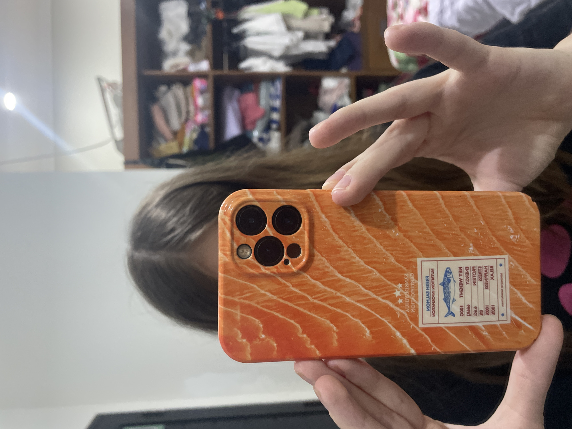 In an indoor setting in Denpasar, Indonesia, a person is holding a smartphone, viewed from the phone's camera perspective. Only the person's hands and a portion of their hair are visible, suggesting they are either taking a selfie or looking into the camera. The focus is on the smartphone, which features a distinctive orange case designed to resemble a cut of salmon fillet.
The phone case incorporates a mock nutritional label, which appears reversed in the image due to the camera's orientation. Visible text, interpreted in its correct orientation, includes "PREMIUM QUALITY", "CERTIFICATION", and "NUTRITION INFORMATION". Under a graphic of a fish, likely salmon or mackerel, phrases like "NET WEIGHT 100g", "ENERGY", "PROTEIN", "FATS", and "SUGAR" are present, along with various numerical values. The word "RELAX" is also visible on the label.
In the background, a wooden shelving unit with multiple compartments is partially visible and slightly out of focus. It contains various items, including what appear to be folded clothes or fabric. A bright light source on the left side of the background creates a noticeable glare. The overall lighting suggests an indoor environment. No specific emotions are discernible.