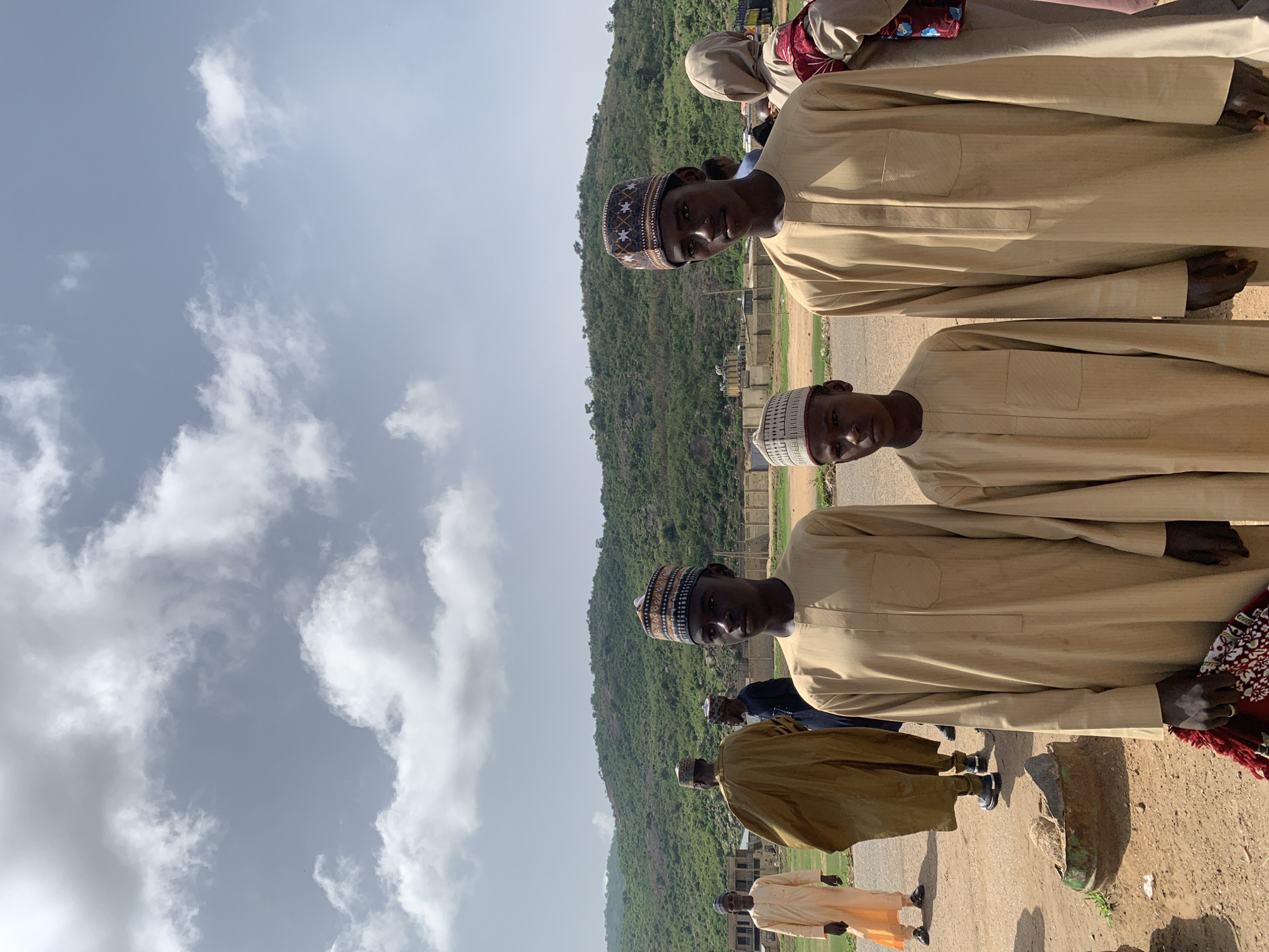 The image shows a group of men standing outdoors in Naraguta Hausawa, Nigeria. They are wearing traditional Hausa attire, including embroidered caps. A large, green hill rises in the background, with a cloudy sky overhead. Some buildings are visible near the base of the hill. The ground is dirt and gravel. The men are standing in a row, facing towards the camera.