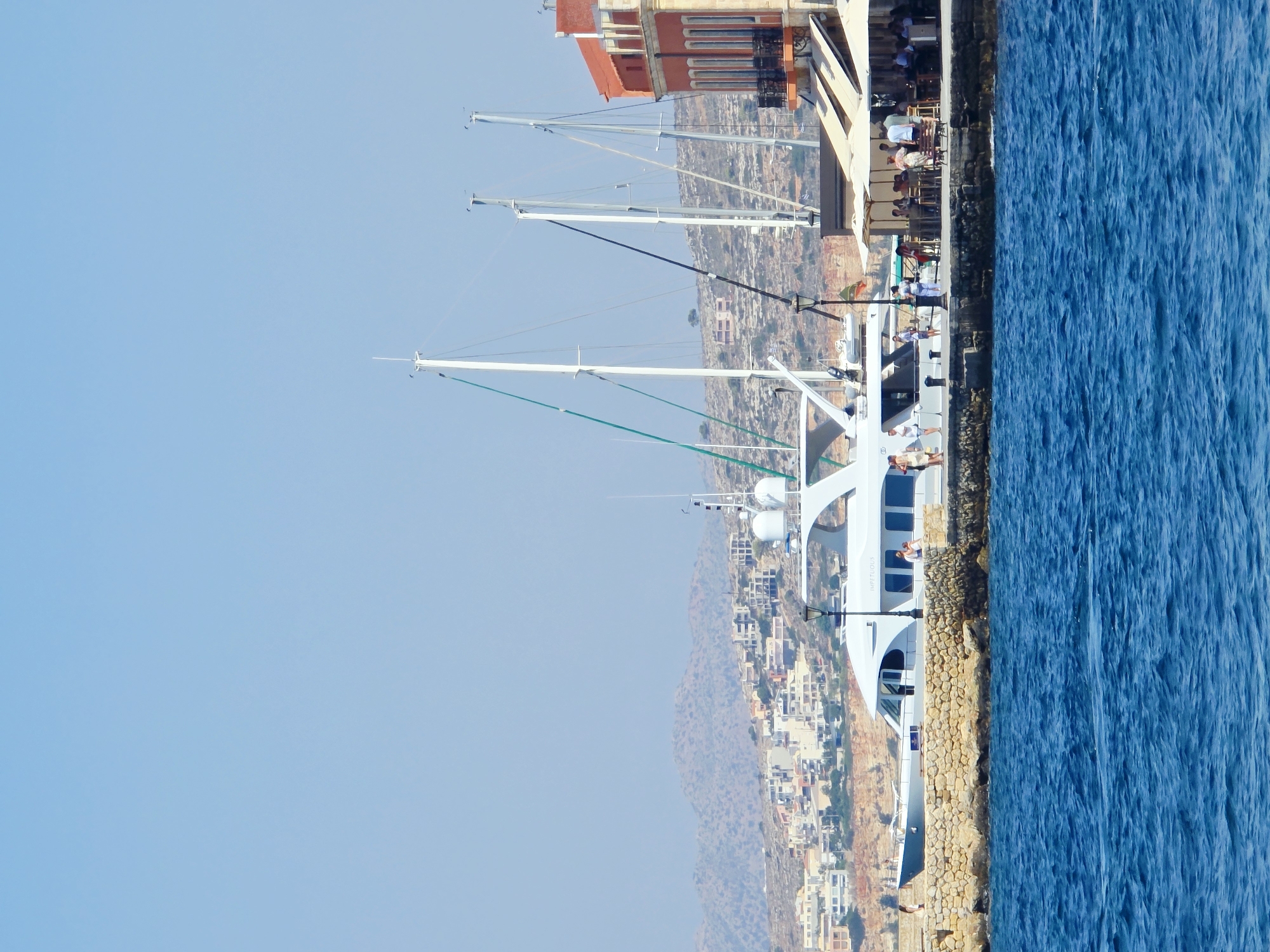 The image depicts a scene in the city of Chania, Greece, likely along its picturesque Venetian harbor. The foreground is dominated by deep blue, rippling water, suggesting a bright and sunny day.
In the midground, a stone quay lines the waterfront, with a large, white motor yacht prominently docked. The yacht has a sleek, modern design with multiple windows. Adjacent to the yacht, a paved walkway is visible, populated by several individuals. Some are strolling, while others appear to be seated at outdoor tables belonging to a cafe or restaurant. The cafe has several white umbrellas providing shade.
Further back, several tall white sailboat masts rise into the clear blue sky, indicating a marina or harbor setting with both motor and sailing vessels. Behind the quay, a cluster of buildings forms the cityscape. The buildings exhibit a mix of architectural styles, with some featuring orange or ochre facades and balconies. Beyond the immediate city, a hillside is visible, dotted with more buildings and sparse vegetation, leading to distant, hazy mountains under the bright sky.
The overall impression is one of a vibrant coastal town, with a blend of leisure activities, maritime presence, and urban architecture under a clear, sunny sky.
