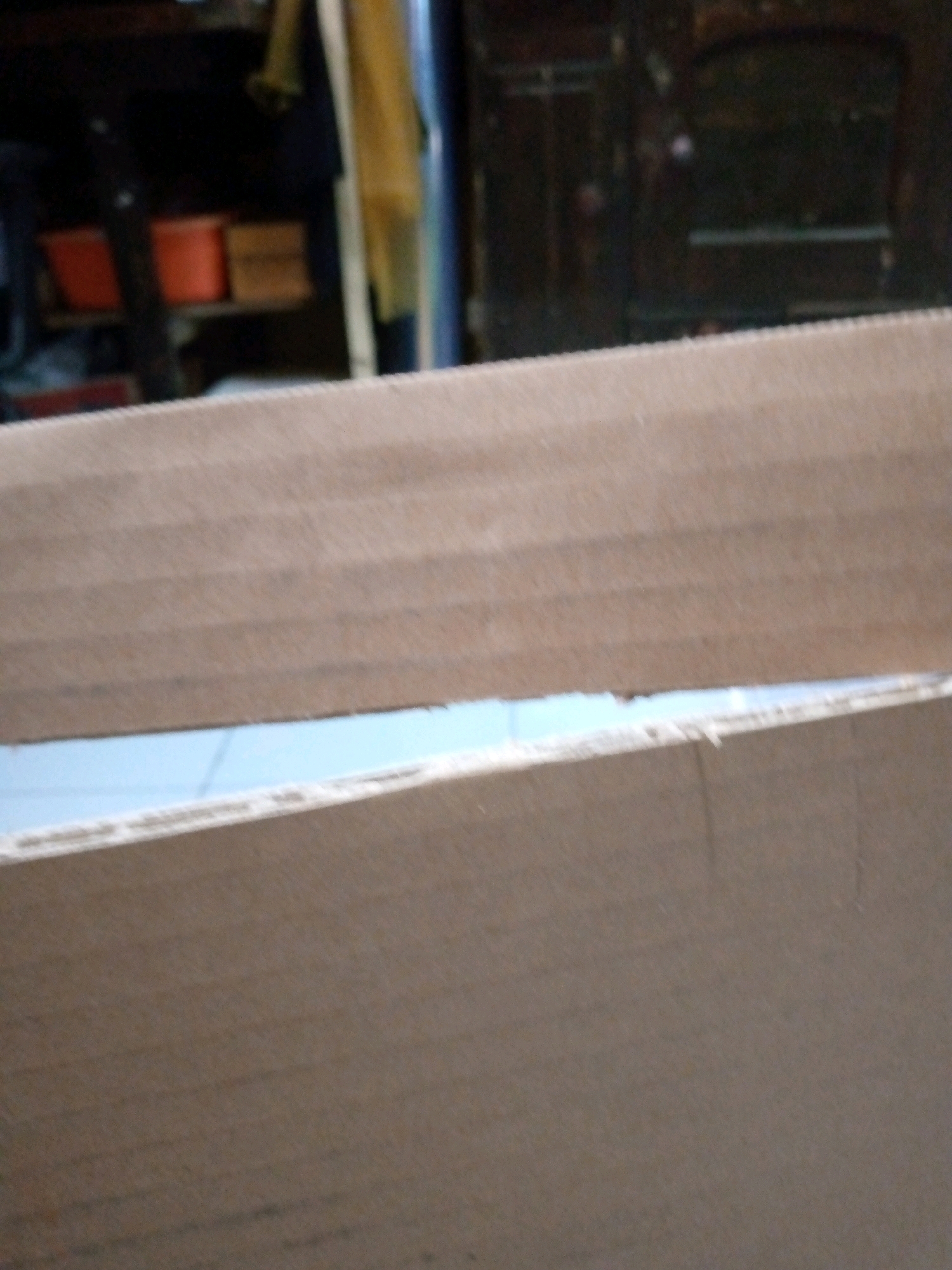 The image shows a close-up view of corrugated cardboard. In the background, out of focus, there appears to be a shelf with two orange buckets and some wooden crates. The environment is indistinct, but the presence of the cardboard suggests a storage or packing area. The scene is indoors, and the lighting is dim. There are no people or animals visible in the image. The location in Sumberwono, Indonesia is not evident from the visual information provided.