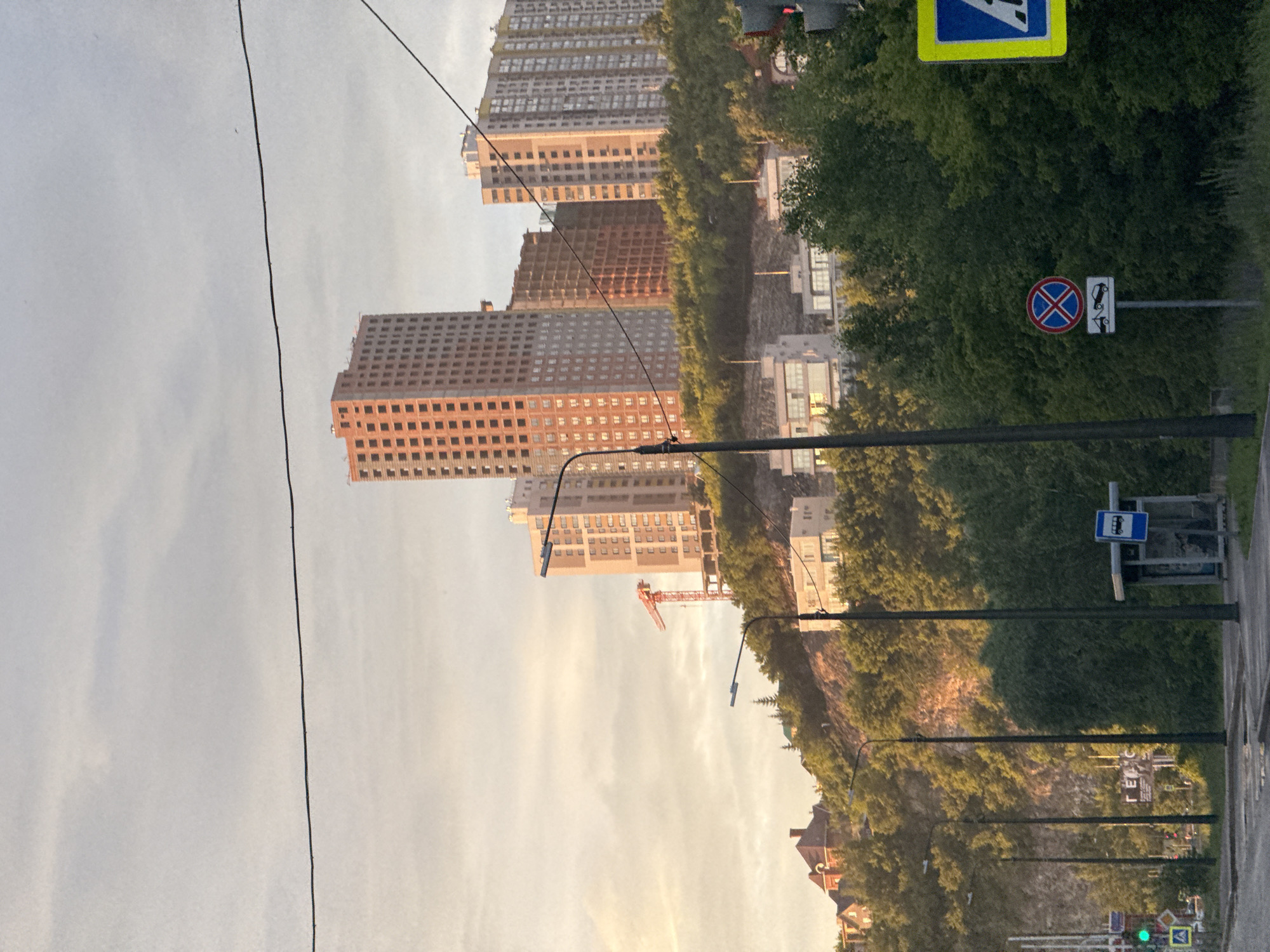 In an urban setting at dusk, a group of unfinished high-rise apartment buildings are visible on a hillside. The buildings are primarily constructed with brick and concrete, with some featuring scaffolding. A red construction crane is also present. In the foreground, a paved road runs alongside a line of trees and streetlights. To the right, there is a bus stop and traffic signs, including a no parking sign with a towing symbol and a pedestrian crossing sign. The sky is overcast with a soft, warm light, indicating either sunrise or sunset.
