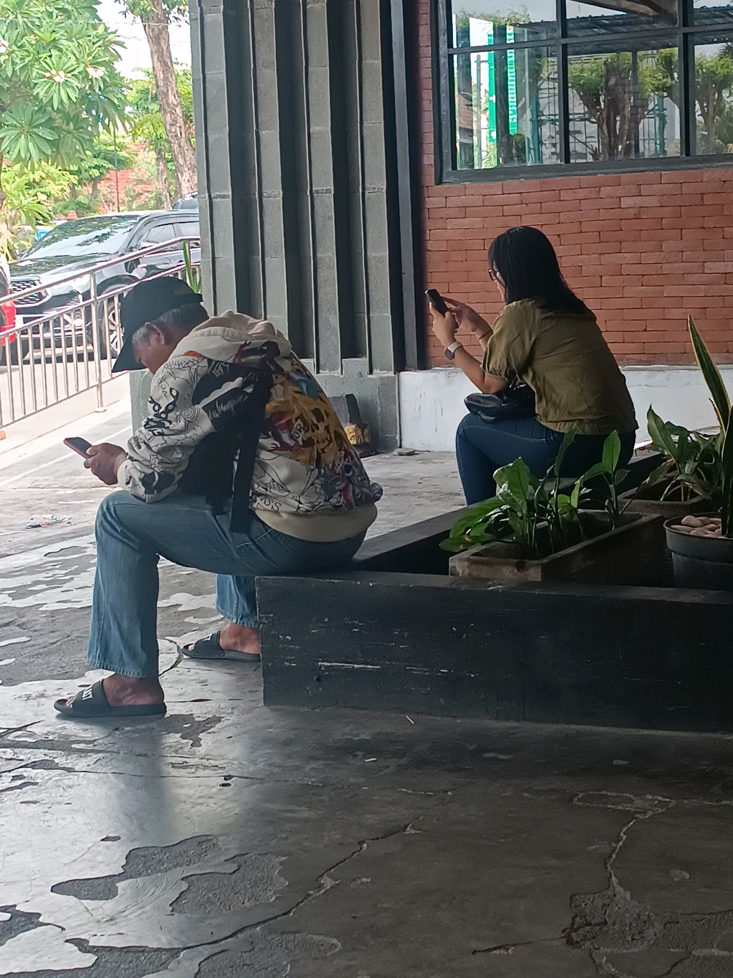 The image shows two people sitting outdoors. On the left, a person wearing a black baseball cap, a colorful jacket, and blue jeans is looking at their phone. They are sitting on a dark, rectangular surface. On the right, a person wearing a green shirt and blue jeans is looking at their phone. This person is sitting on the same dark surface. In the background, there is a brick wall, a large window with reflections, and some greenery. The ground is concrete with some cracks and stains. The scene appears to be in a public outdoor area, possibly a cafe or waiting area, located in Denpasar, Indonesia.