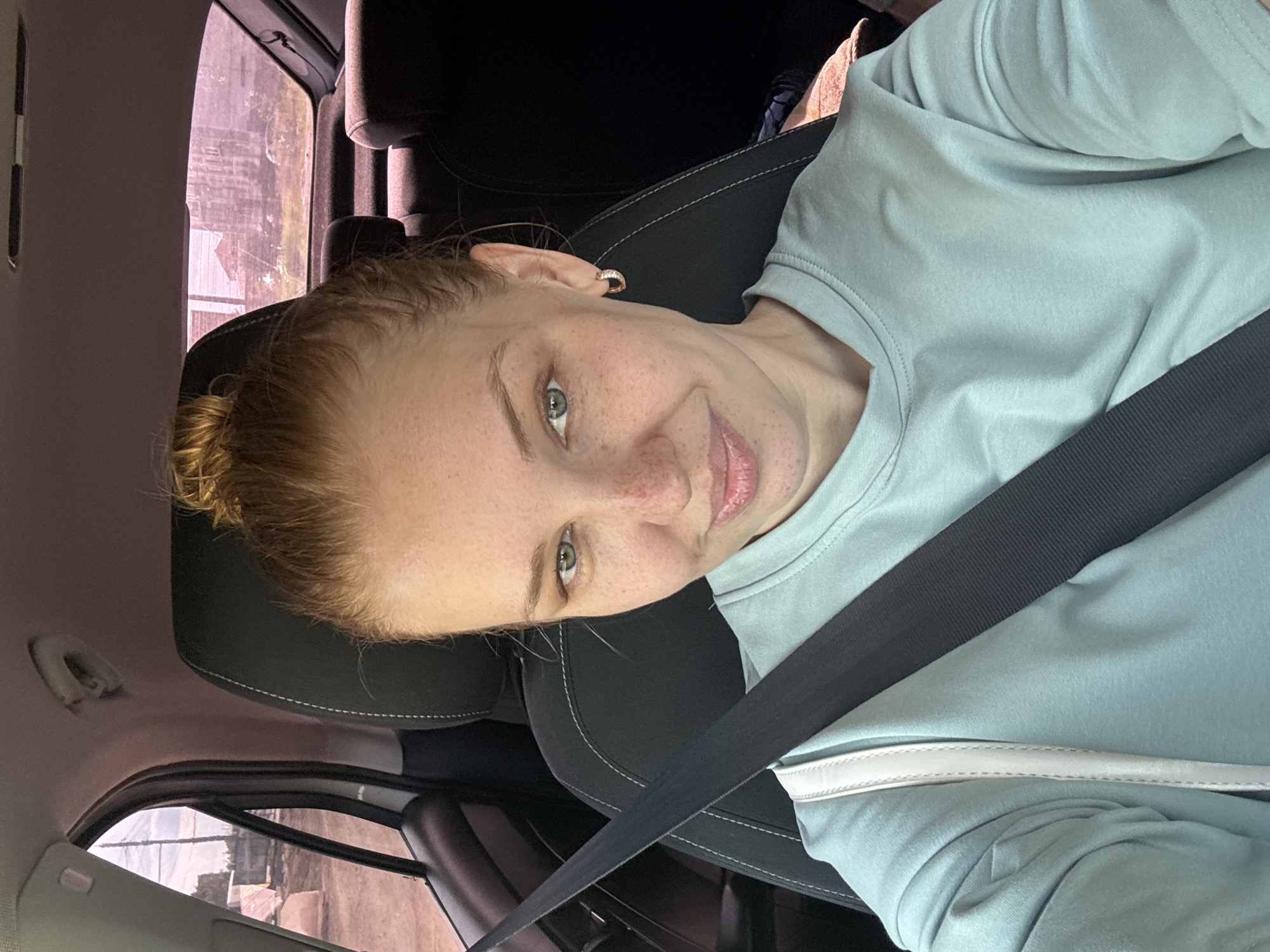 A young woman with blonde hair tied up in a bun and light green eyes is sitting in the passenger seat of a car. She is wearing a light teal t-shirt and a seatbelt is across her chest. The interior of the car is visible, including the headrest of the seat and part of the dashboard. Through the car window, a hazy outdoor scene can be seen, with some buildings and vegetation in the background, suggesting an urban or suburban environment, potentially in or around Rusanovo, Russia. The overall lighting suggests it is daytime.