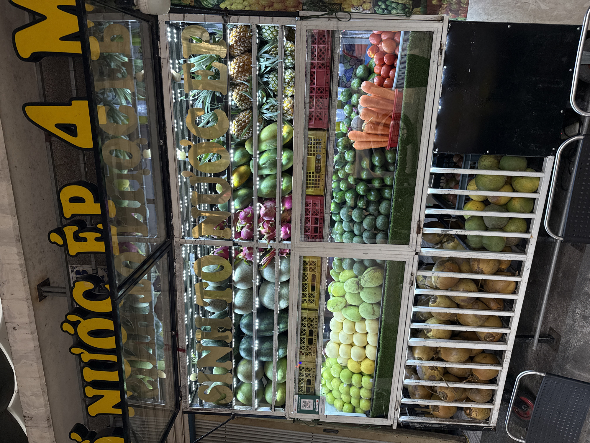 The image depicts an outdoor fruit juice stand in Da Nang, Vietnam. The stand is well-lit and features a variety of fresh fruits displayed in refrigerated compartments. Prominent signage in yellow and black, with Vietnamese characters, indicates "SINH TO NUOC EP" and "4 M", which translates to "Smoothie Juice" and likely refers to prices or special offers.
In the foreground, a close-up shows rows of coconuts displayed in a caged section of the stand. To the right, a black panel is visible. In the background, various fruits are arranged on shelves, including what appear to be watermelons, melons, dragon fruit, pineapples, avocados, carrots, and tomatoes. The overall scene suggests a bustling urban environment, common for street-side vendors in Da Nang. No people are clearly visible in the frame.