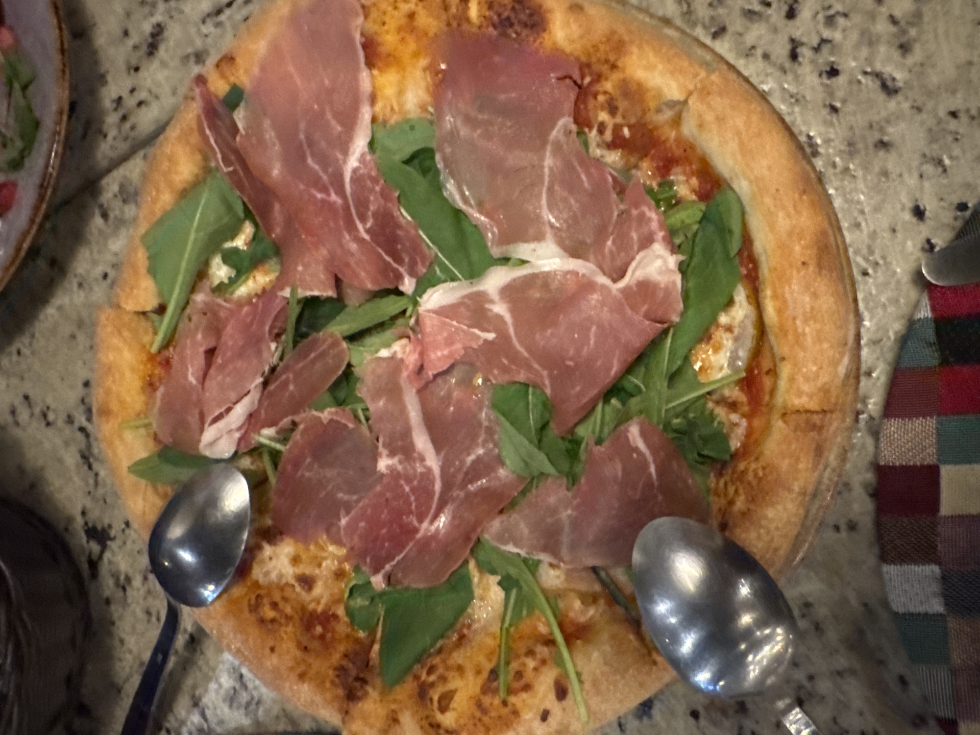 A high-angle shot shows a pizza topped with arugula and prosciutto. Two spoons are positioned on either side of the pizza. A plaid cloth napkin rests on the right, while a blurred dish is visible on the left. The surface beneath is a speckled gray. The image provides no indications regarding the location in the city of Puebla, Mexico.