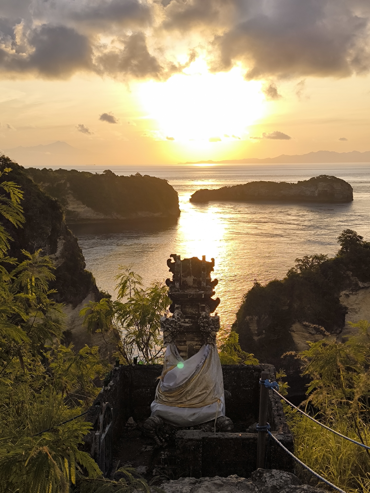 The image captures a sunset view at Nusa Penida, Indonesia, overlooking the ocean and offshore islands. In the foreground stands a stone Balinese shrine adorned with a white and gold cloth, positioned on a stone platform. The scene is framed by foliage on either side. The ocean reflects the golden light of the setting sun, creating a shimmering effect. Several islands dot the horizon under a partly cloudy sky, with the sun obscured by clouds. A wooden post with blue ropes functions as a safety barrier near the shrine.