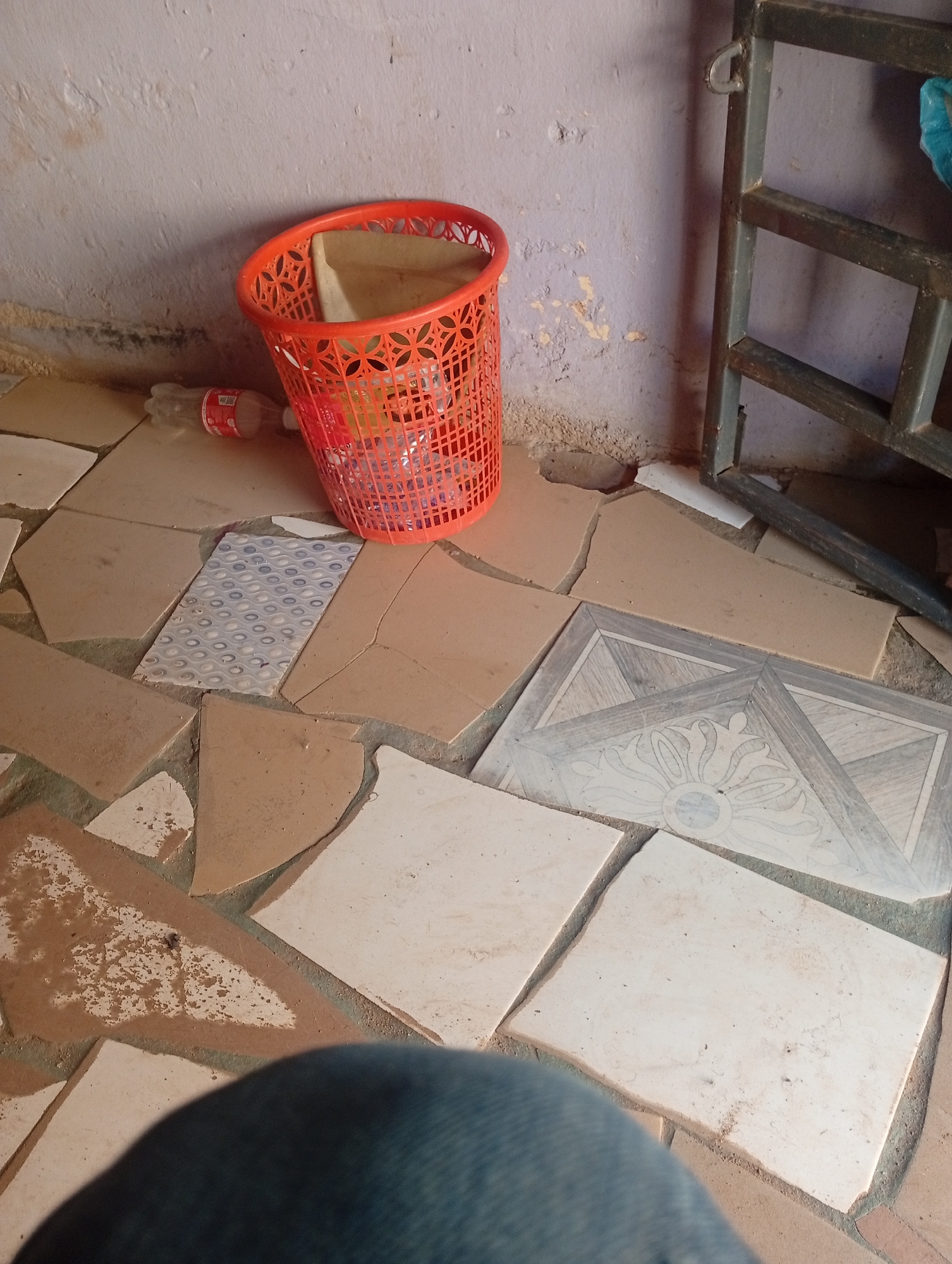 The image depicts an interior scene, likely in Rafin Par, Nigeria. A bright orange plastic waste bin, filled with what appears to be paper or plastic waste, is situated near a light purple, damaged wall. To the left of the bin, a partially consumed bottle of Coca-Cola lies on the floor. The floor itself is an uneven mosaic of broken tiles in various shades of beige and white, with one small, patterned tile in blue and white visible. In the background, a dark metal structure, possibly part of a gate or shelving unit, stands against the wall. The overall environment appears to be somewhat neglected, with debris and cracked tiles suggesting a state of disrepair. A portion of a person's denim-clad leg is visible in the foreground, implying their presence at the scene.