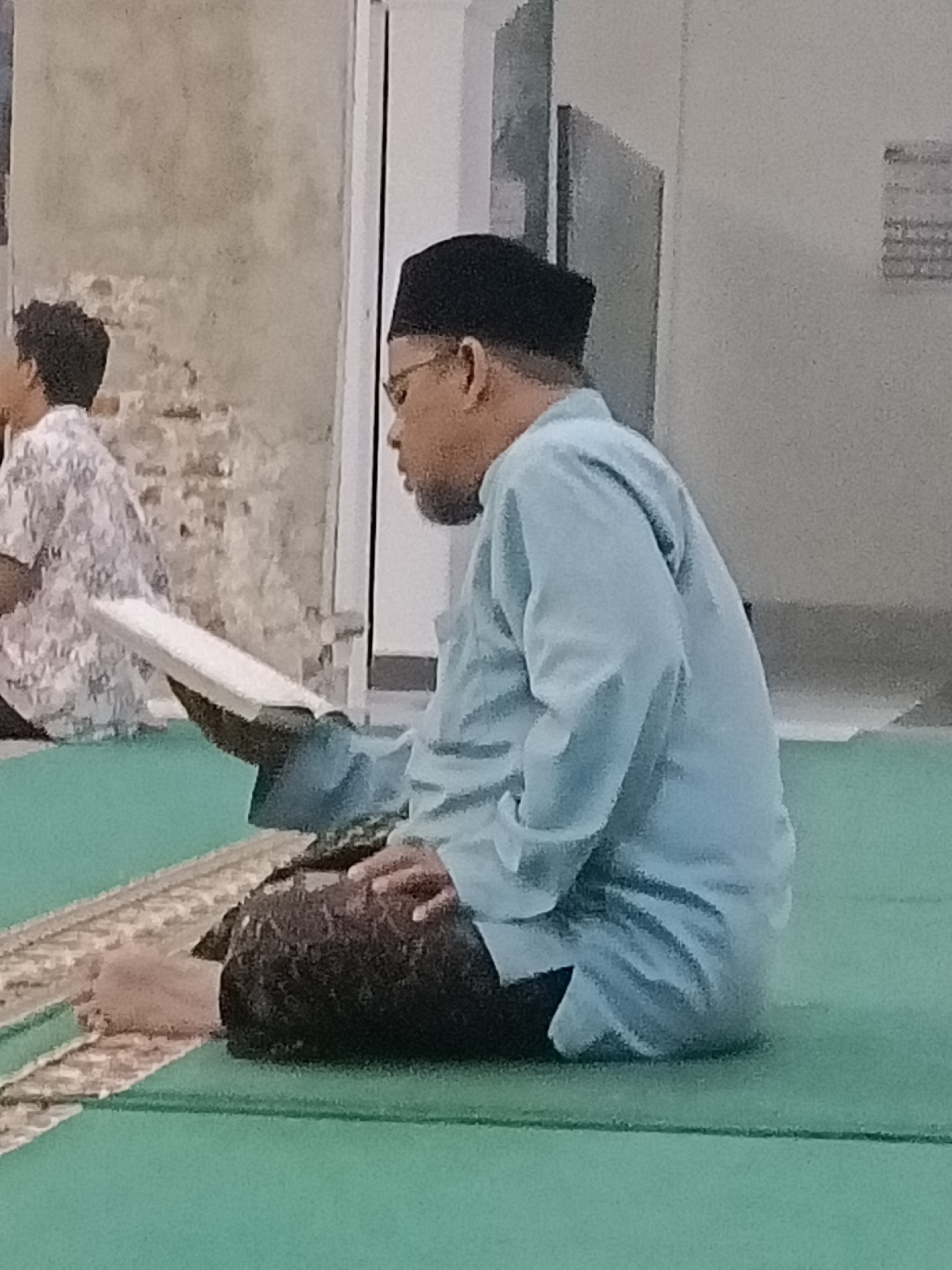 The image depicts two men in what appears to be a prayer room. The man in the foreground, on the right, is wearing a light blue tunic and a dark cap, and is sitting on a green carpet, holding a book. His head is turned to his left, and he is looking down at the book. His right leg is crossed in front of his left leg. The man in the background, on the left, is wearing a patterned shirt and also appears to be looking down. The environment suggests an indoor setting with plain walls and a green carpet. The lighting is somewhat dim, possibly indicating evening or an overcast day. The image quality is moderate, with some pixelation visible. The context of the men wearing religious attire and the presence of a book suggests a religious gathering or prayer session. It is not possible to determine the specific location within Kediri City, Indonesia, from the visual information alone.