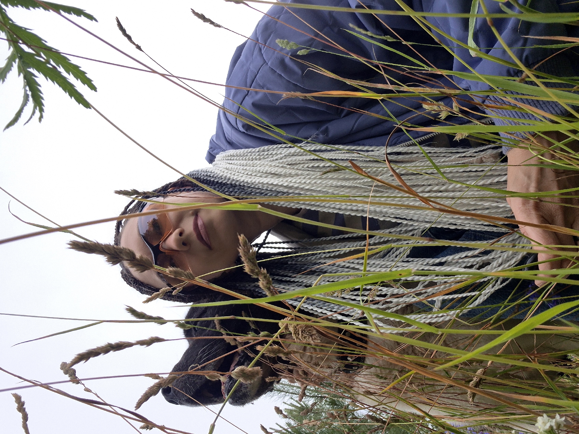 An outdoor, eye-level, medium shot captures a person and a dog amidst tall grass. The person, positioned on the right and partially obscured by foliage, wears a blue quilted jacket, sunglasses with orange lenses, and has light-colored braided hair. Their face is turned towards the camera. To the left, a black dog with a tan chest is visible, its head and snout facing upwards and to the left, its mouth slightly open. The environment suggests a natural setting, likely a field or park, with the sky appearing overcast and pale. The overall composition is framed by numerous blades of grass in the foreground, creating a sense of depth and intimacy with the subjects. The location is identified as Chaikovsky, Russia.