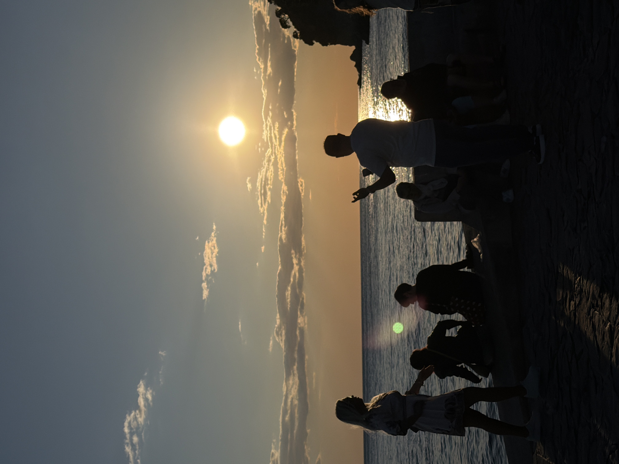 This photograph captures a scenic coastal moment, likely during sunset or sunrise, in Petrovac centar, Montenegro. The bright, circular sun is prominently featured on the left, casting a radiant golden-yellow light across the sky. The sky transitions from a darker blue-grey on the far left to warm orange and yellow hues around the sun, with a distinct, elongated cloud formation stretching vertically and catching the light.
In the foreground and midground, several people are silhouetted against the luminous sky and water. They are gathered on a stone walkway or promenade that runs along the water's edge. Some individuals are standing, while others appear to be seated or crouching, their forms entirely dark due to the intense backlighting. One person in the midground seems to be gesturing with an open hand. The water, a dark blue, reflects the sun's brilliance with shimmering streaks of light. The overall atmosphere is one of peaceful observation, as the figures gaze out towards the horizon. No text is visible in the image.