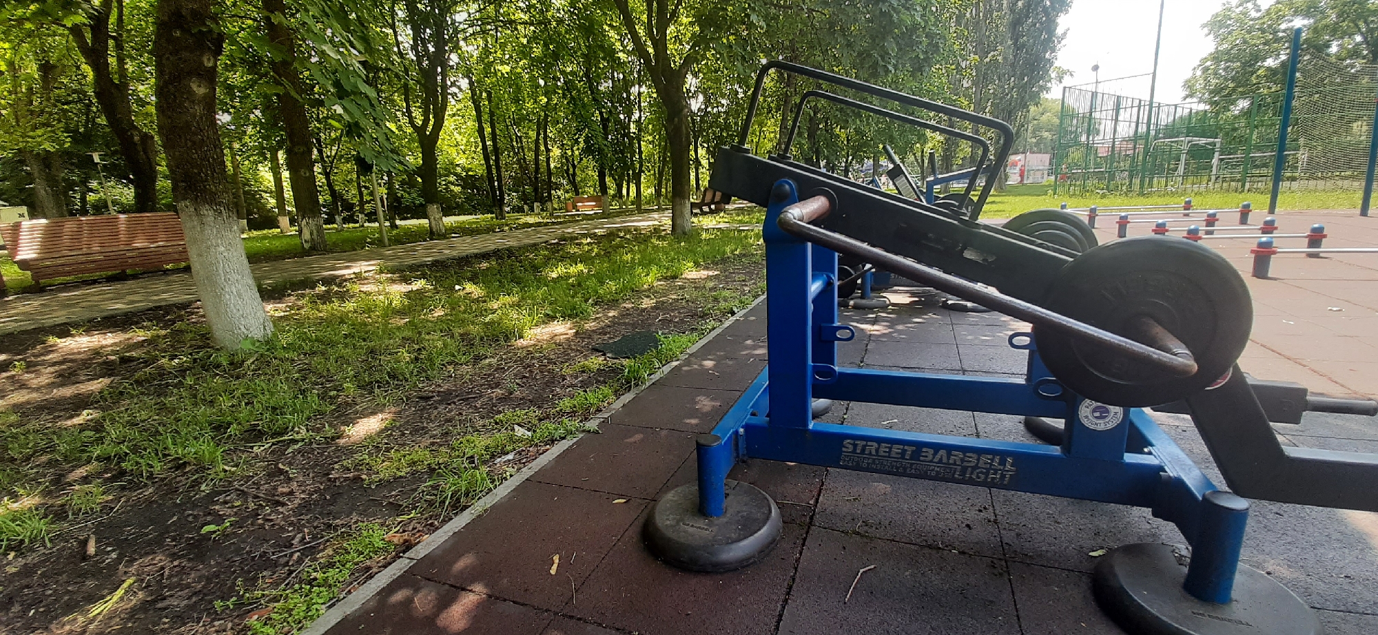 The image shows an outdoor exercise machine in a park, likely located in Yessentukskaya, Russia. The machine is blue and black and appears to be a weight-lifting apparatus with at least one large weight plate visible. It is situated on a dark rubberized surface, which is bordered by a paved pathway. To the left of the pathway, there is a wooden bench and grassy areas with trees. In the background, there is a fenced sports field, possibly for soccer or basketball, with more trees and a bright sky. No people are visible in the scene.
