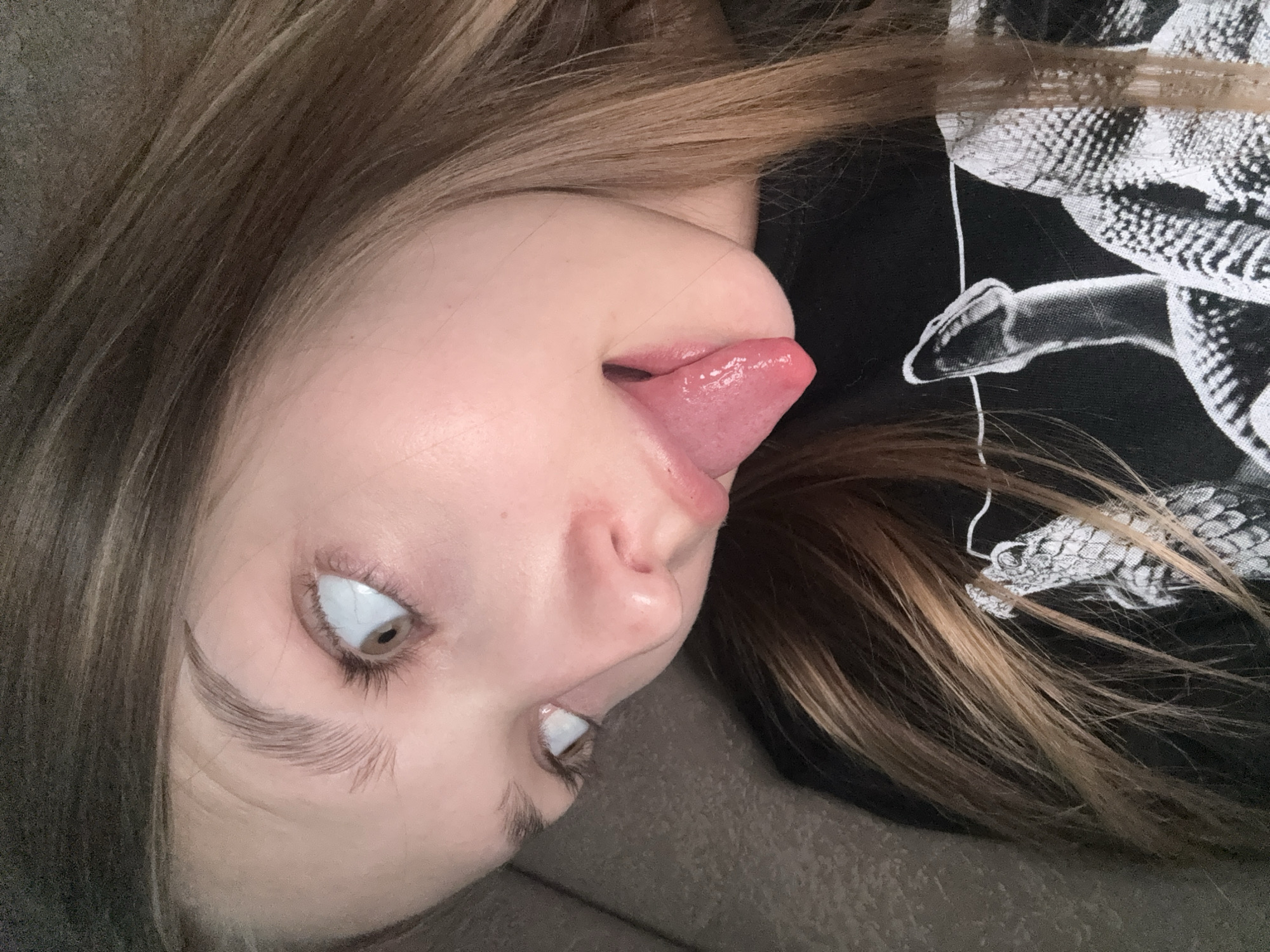 A woman is lying on a brown couch. Her head is tilted to the left, and she is sticking her tongue out. She has brown hair and brown eyes. She is wearing a black shirt with a white snake design. The image was taken indoors. There is no indication of the location being Марковский, Russia.